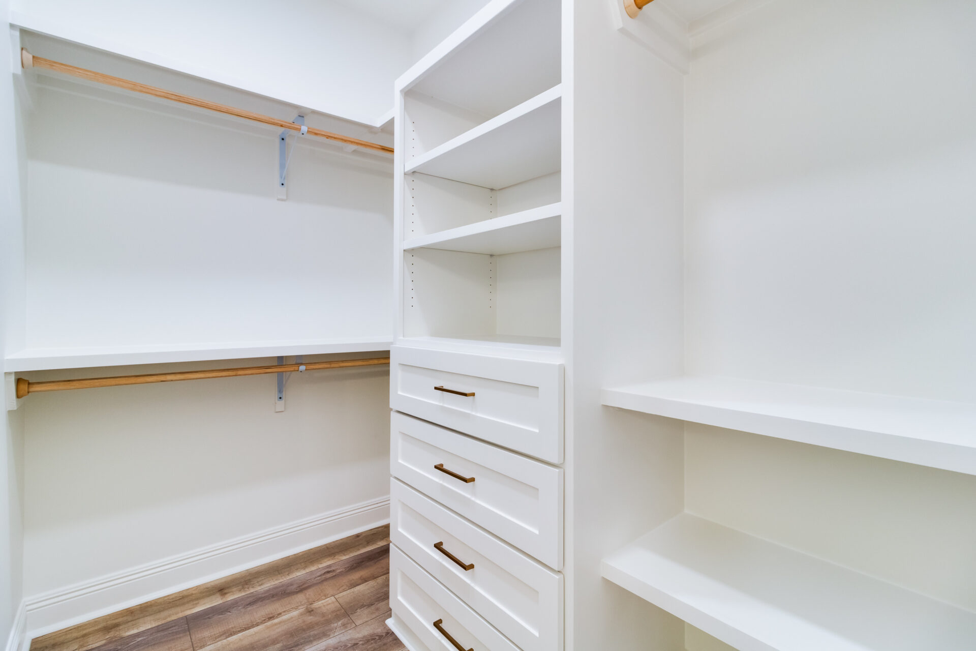 Spacious walk-in closet with white shelves and drawers, expertly crafted by Hebert Reno. The closet features wooden rods for hanging clothes and open shelves for storage. The flooring is light wood, and the overall decor is bright and minimalistic.