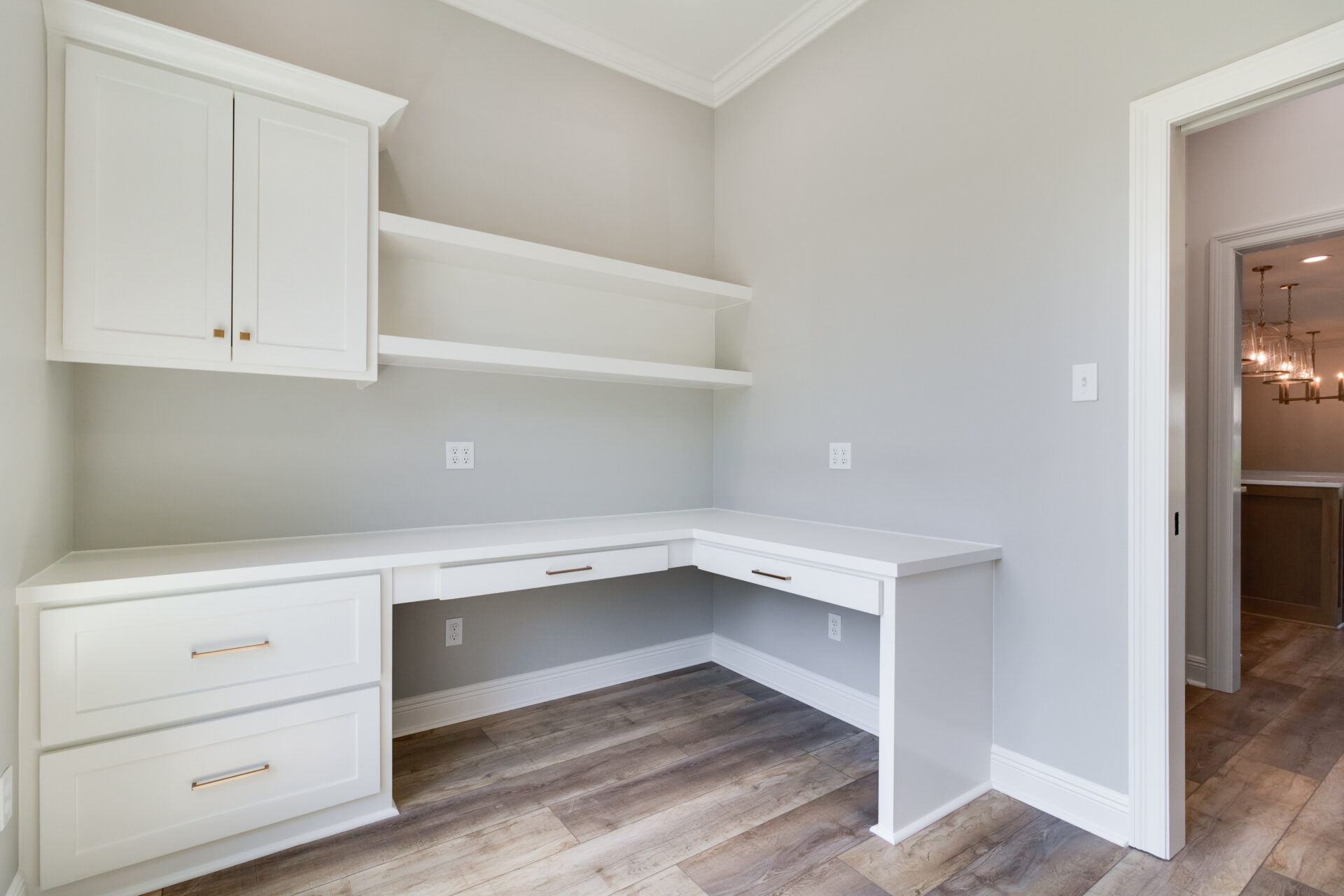 A home office with an L-shaped white Hebert desk featuring brass handles, overhead cabinets, and open shelves. The room has light gray walls, wooden flooring, a white door, and visible light fixtures in an adjoining Reno room.