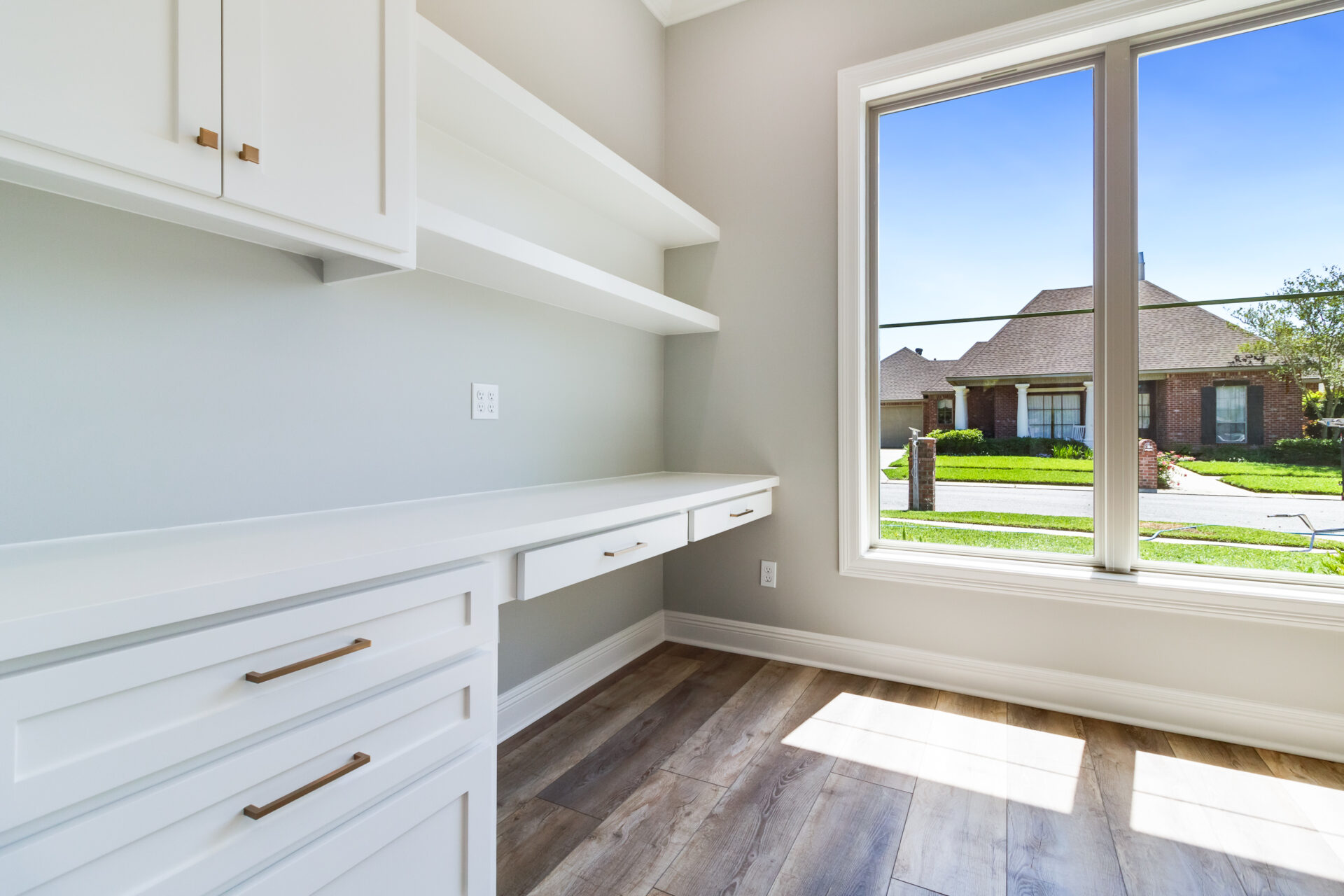 Bright room with white cabinets and shelves built along a wall, featuring a large window that offers a street view of a suburban neighborhood. The room, enhanced by Hebert Reno, has light gray walls and wood-style flooring.