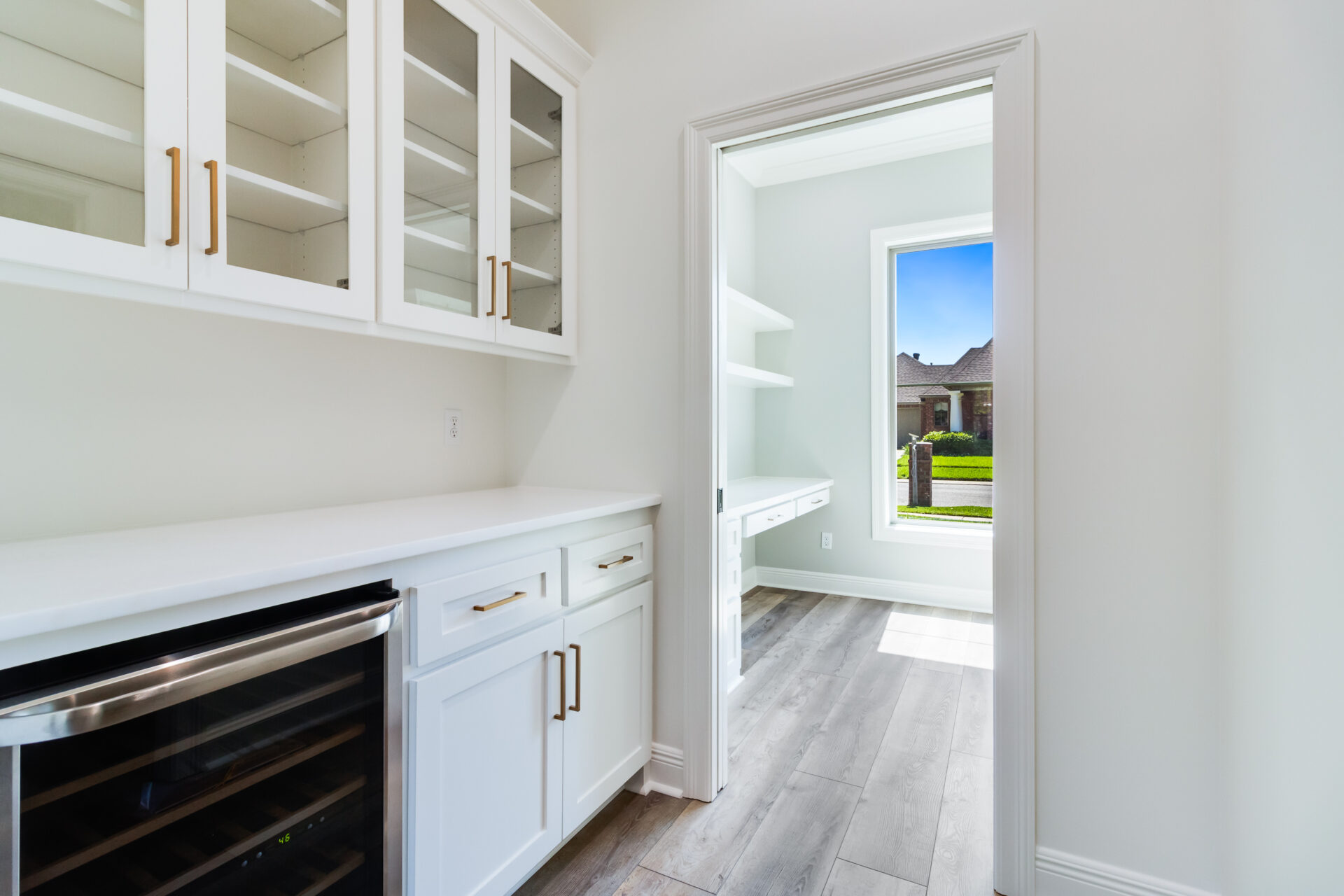 A bright pantry with white cabinets and gold handles, featuring multiple shelves and drawers by Hebert Reno, adjacent to a small office or workspace area with a window facing a grassy outdoor view. The floor is light gray wood, and there is a wine cooler under the counter.