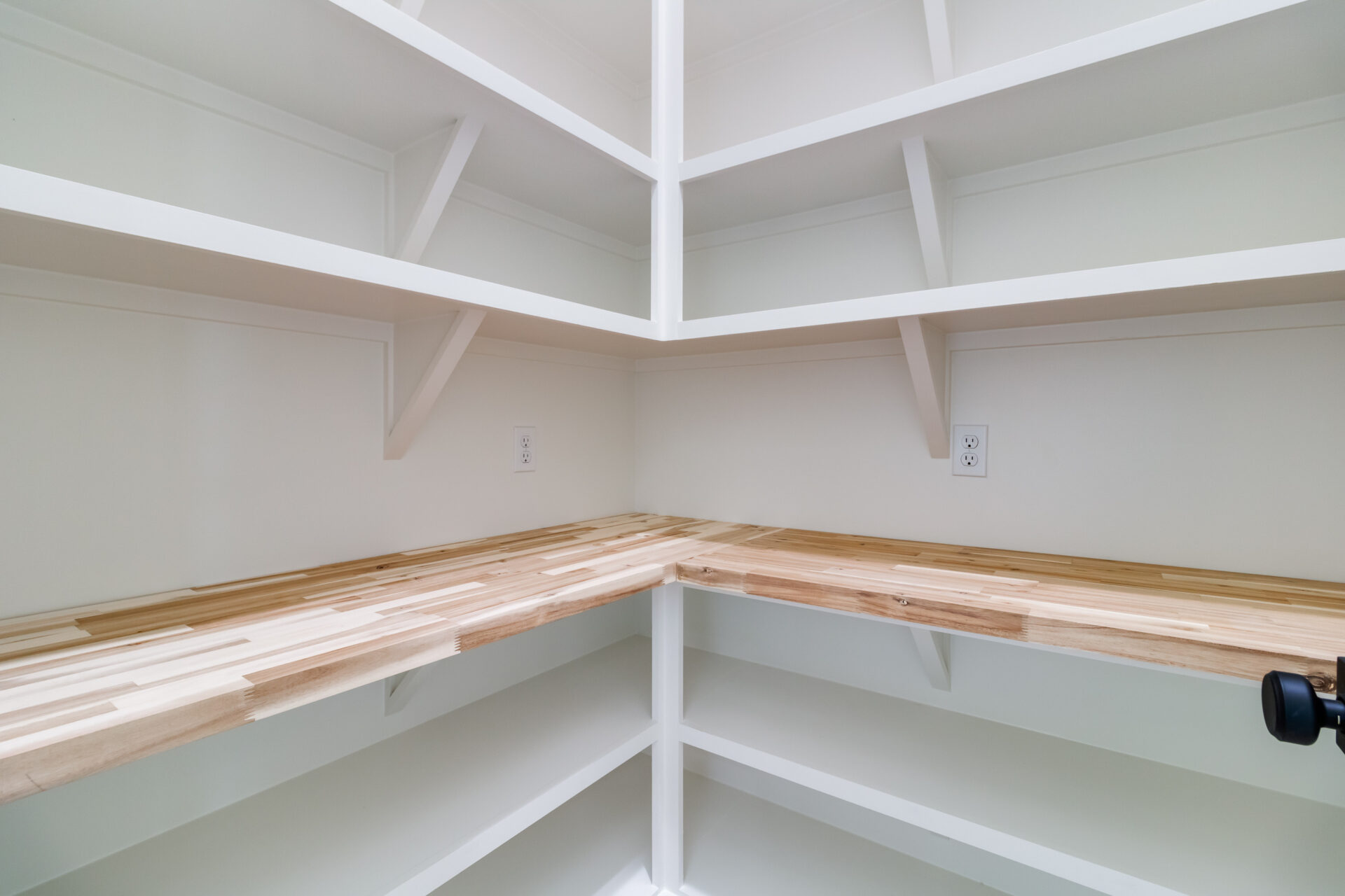 A corner pantry by Hebert Reno features white shelving and a light wood countertop. The empty shelves are complemented by two electrical outlets on the walls above the countertop. This well-lit space, with its white walls and angled support brackets, exemplifies functional elegance.
