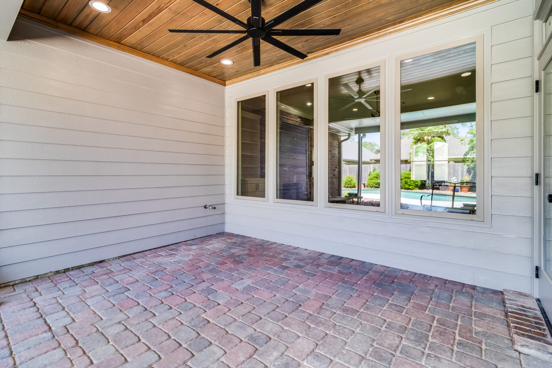 A covered outdoor patio area with brick flooring, white siding on the walls, and a wood-paneled ceiling featuring ceiling lights and a large black ceiling fan. The Hebert Reno patio overlooks a pool through a set of large windows.