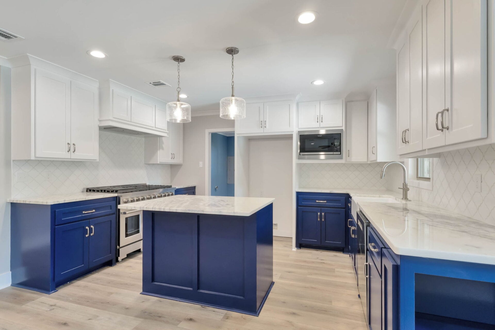 Modern kitchen with white upper cabinets, blue lower cabinets and an island, stainless steel appliances, marble countertops, light wood flooring, and two pendant lights hanging above the island.