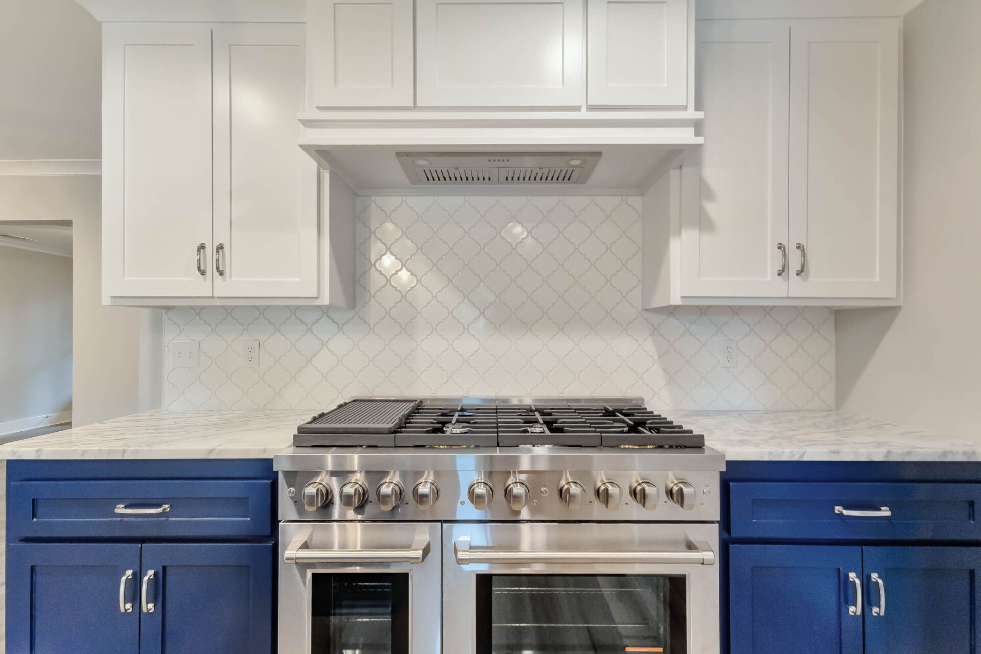 A modern kitchen features a stainless steel gas range with dual ovens, blue lower cabinets, white upper cabinets, and a white tile backsplash with a diamond pattern.