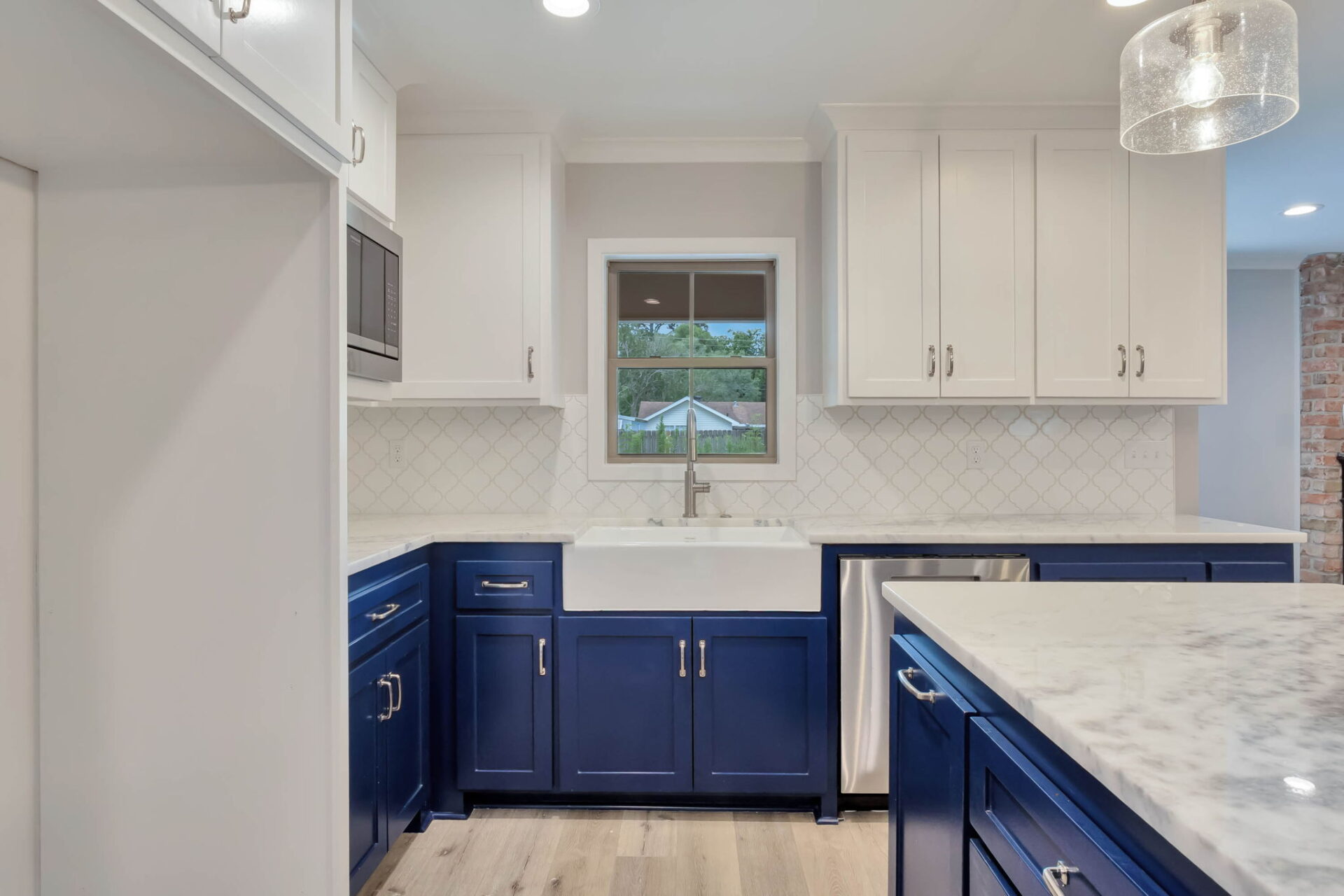 Modern kitchen with white upper cabinets and navy blue lower cabinets, a farmhouse sink under a window, a marble countertop, and a white tile backsplash. A brick wall is partially visible on the right.