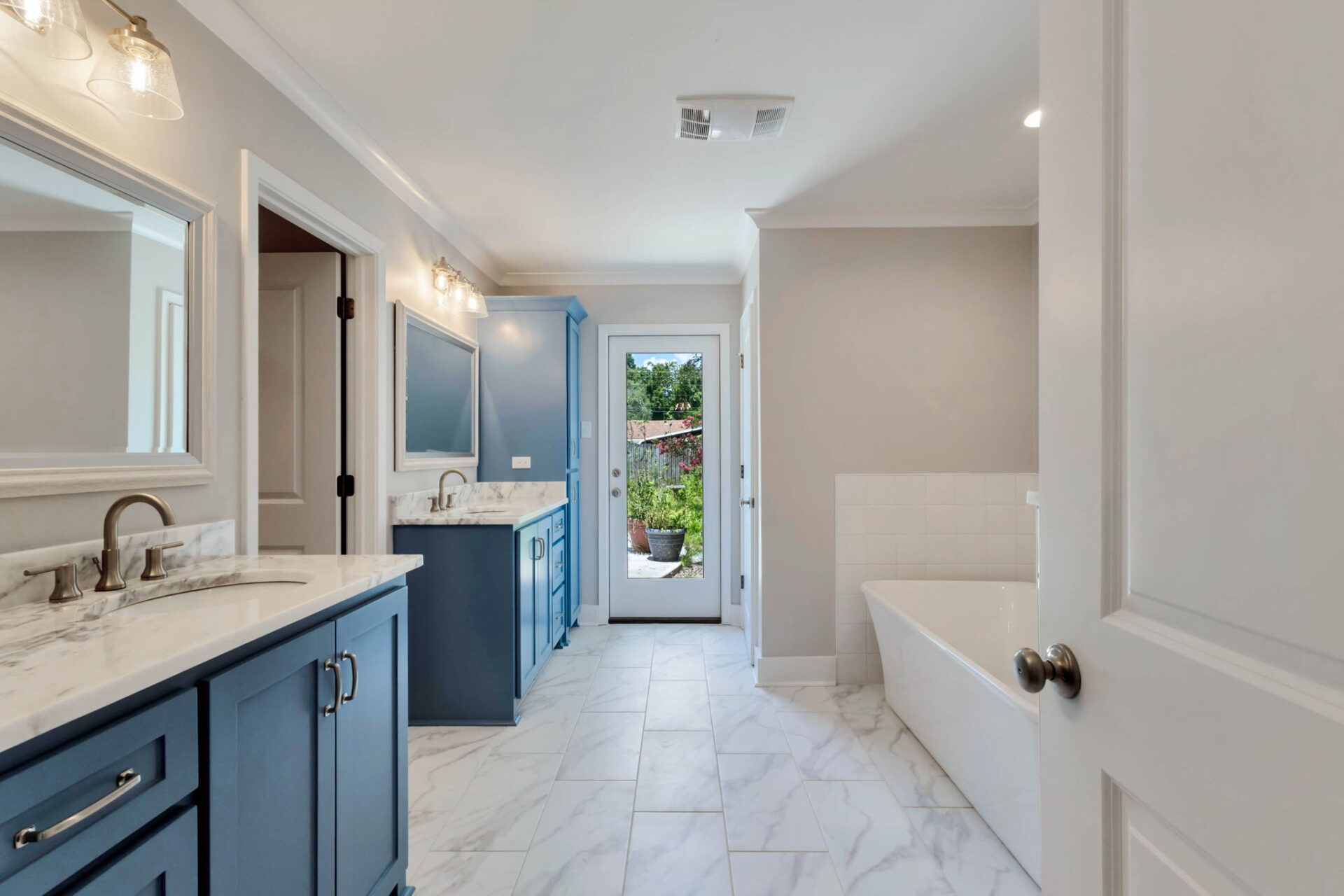 A modern bathroom with blue cabinets, white marble countertops, double sinks, a large mirror, a bathtub, tile floors, and a door leading outside to a garden area.