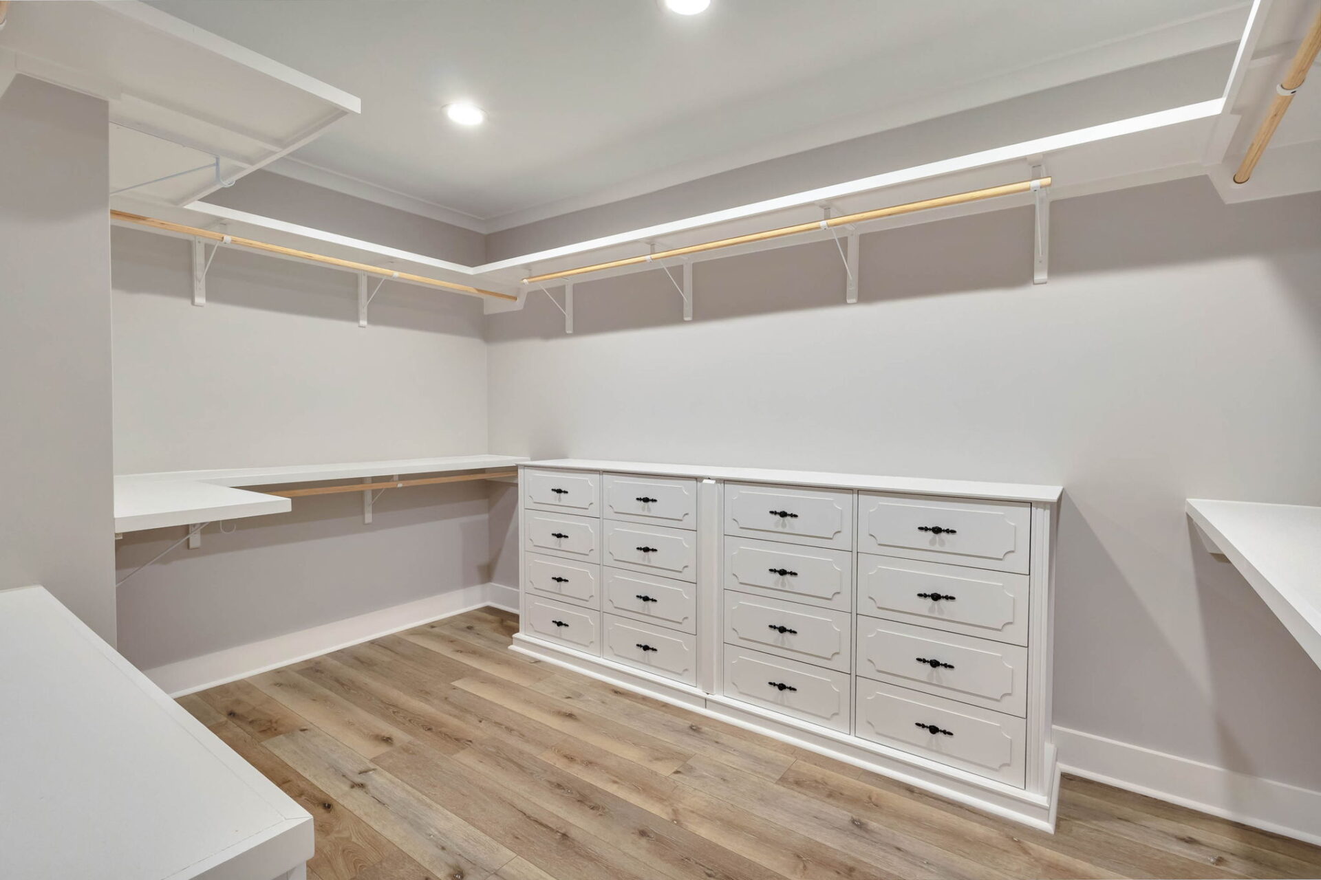 A spacious walk-in closet with white shelves, wooden hanging rods, and two sets of white drawers. The closet has hardwood floors and is brightly lit by recessed ceiling lights.