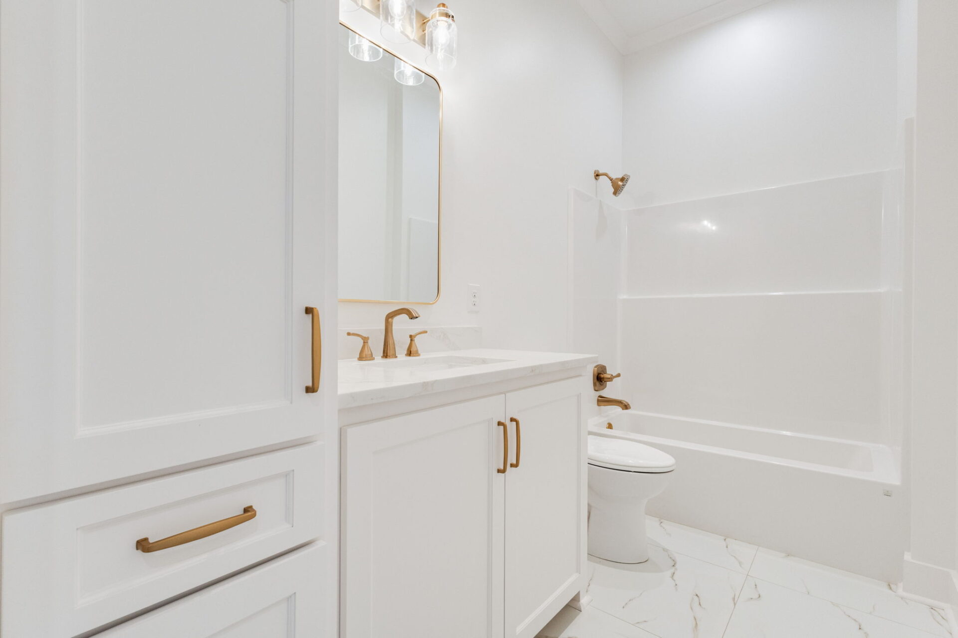 A bright bathroom with white cabinets, gold hardware, a white countertop with dual sinks, a toilet, and a bathtub with a shower, all set against white walls and marble-patterned floor tiles.