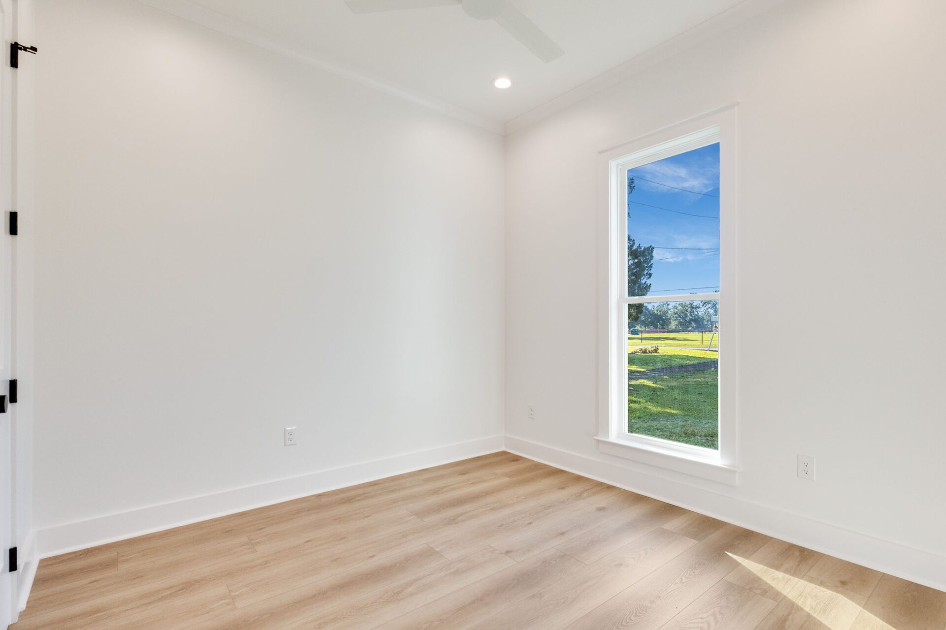 A bright, empty room with white walls, light wood flooring, a large window showing a green lawn and trees outside, white trim, and ceiling lights.