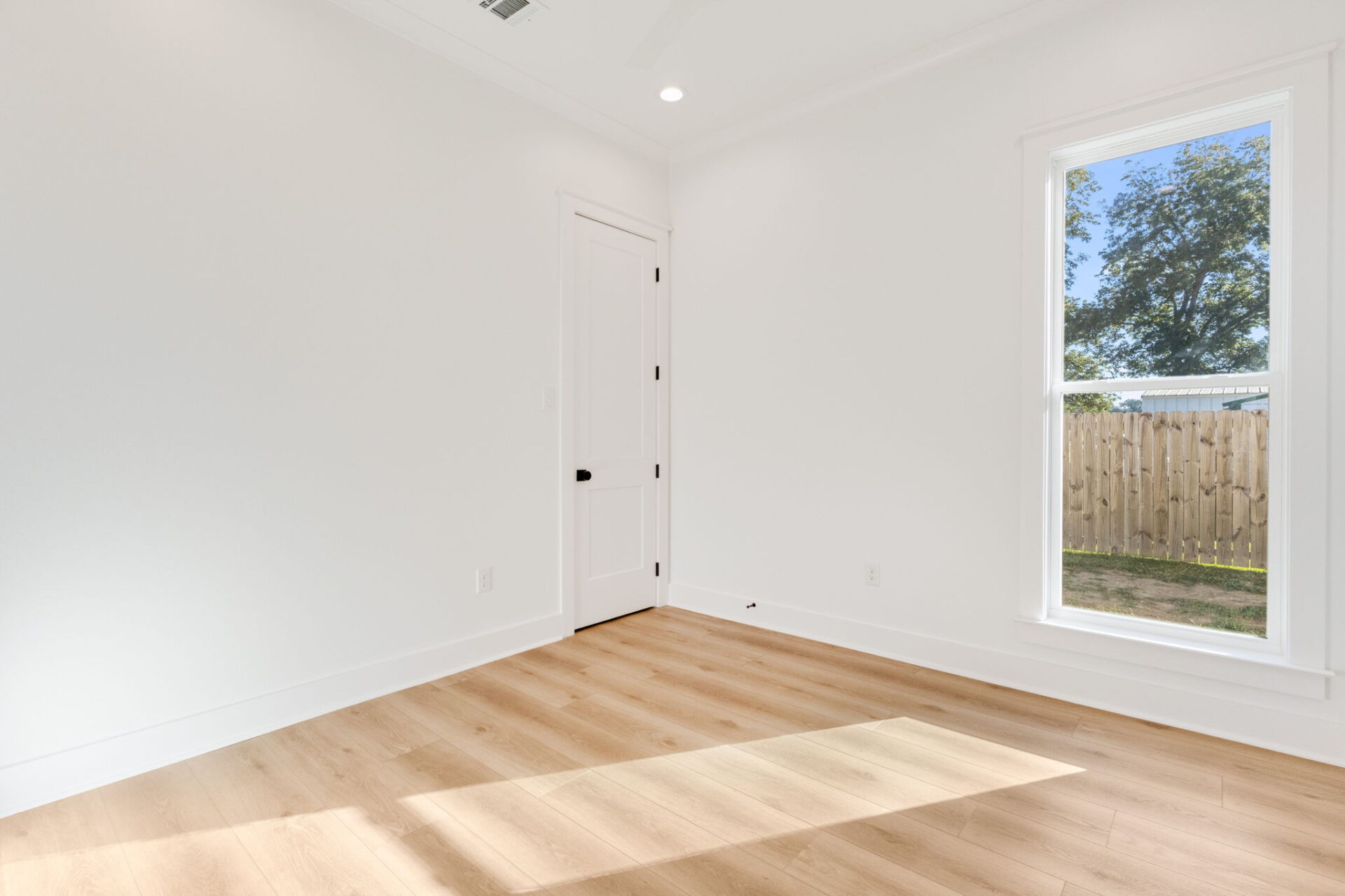 A small, empty room with white walls, light-colored wood flooring, a closed white door with a black handle, and a large window showing a wooden fence and tree outside. Sunlight is streaming in through the window.