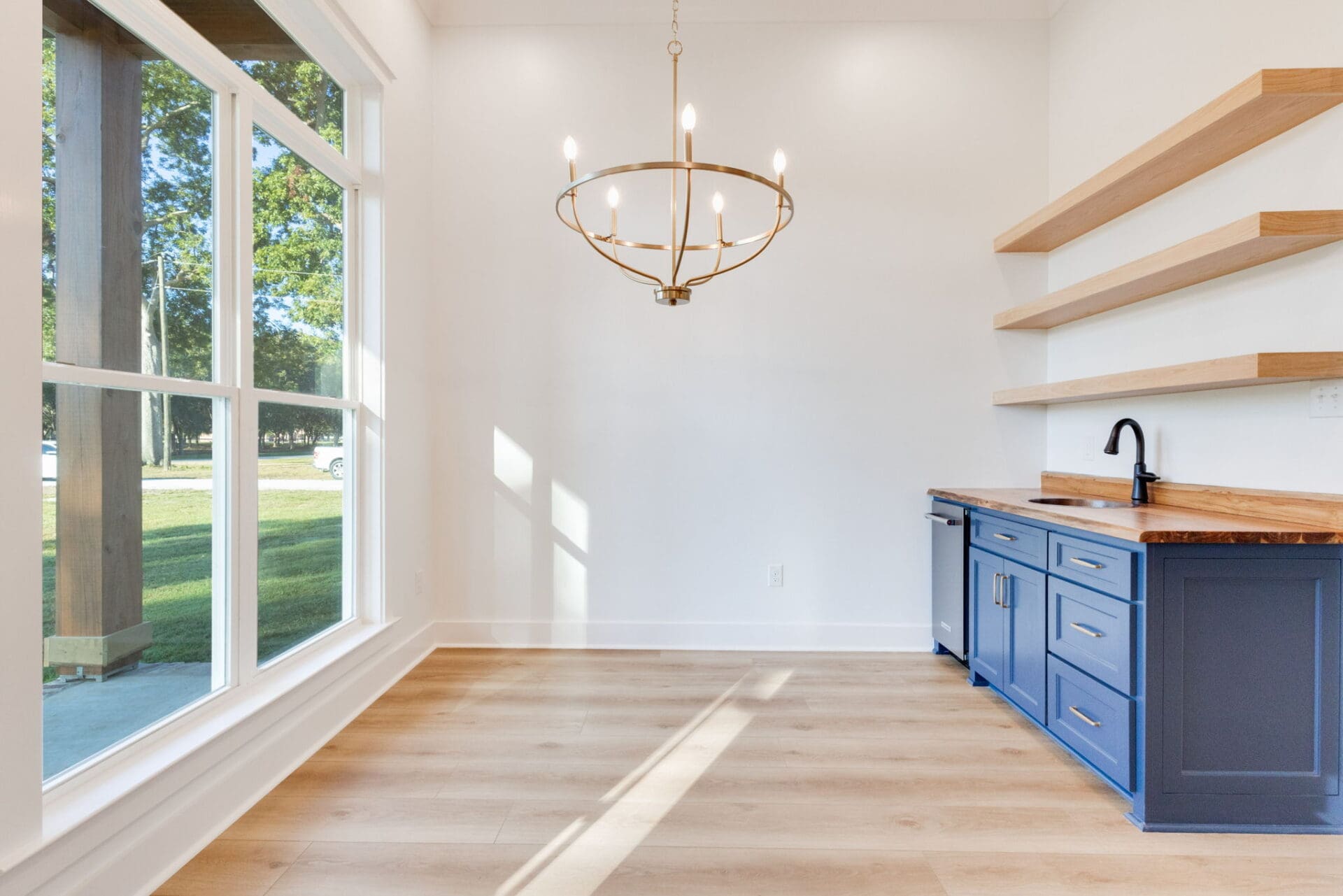 A bright room with large windows, a modern gold chandelier, blue cabinets with wood countertops, open wood shelves, a black faucet, and light wood flooring. The walls are white and the space is empty.