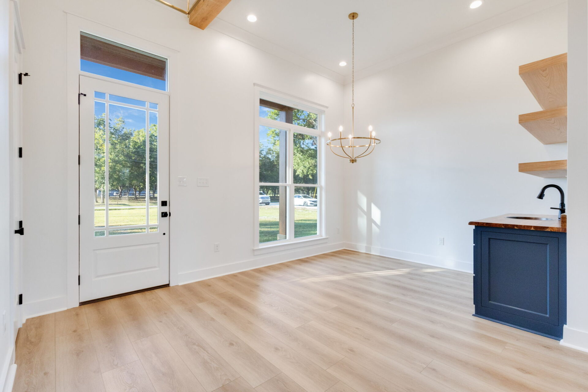 A bright, empty room with light wood floors, white walls, large windows, a glass door, a modern chandelier, open wood shelves, and a small kitchenette with a dark blue cabinet and a sink.