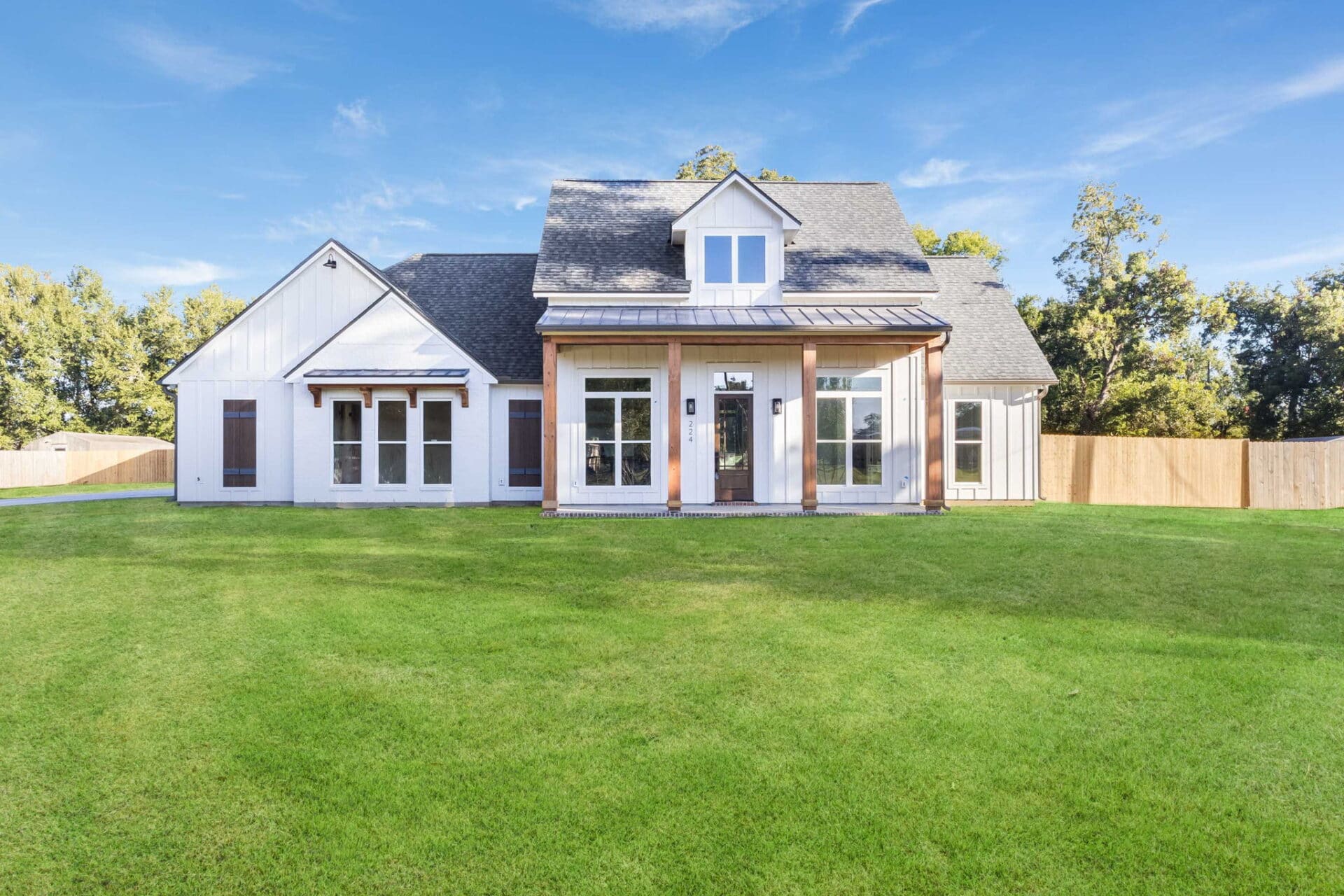A modern single-story house with white exterior walls, large windows, wooden pillars at the entrance, and a manicured green lawn, set against a backdrop of trees under a blue sky.
