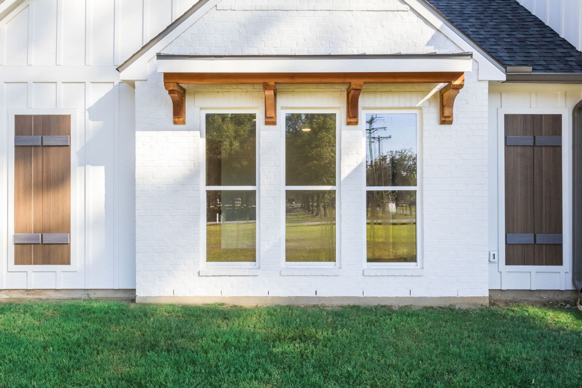 Three tall windows are set in a white brick wall, with a wooden awning above them. The wall has vertical siding on each side, each section featuring a brown shutter. A grassy lawn is in front of the house.
