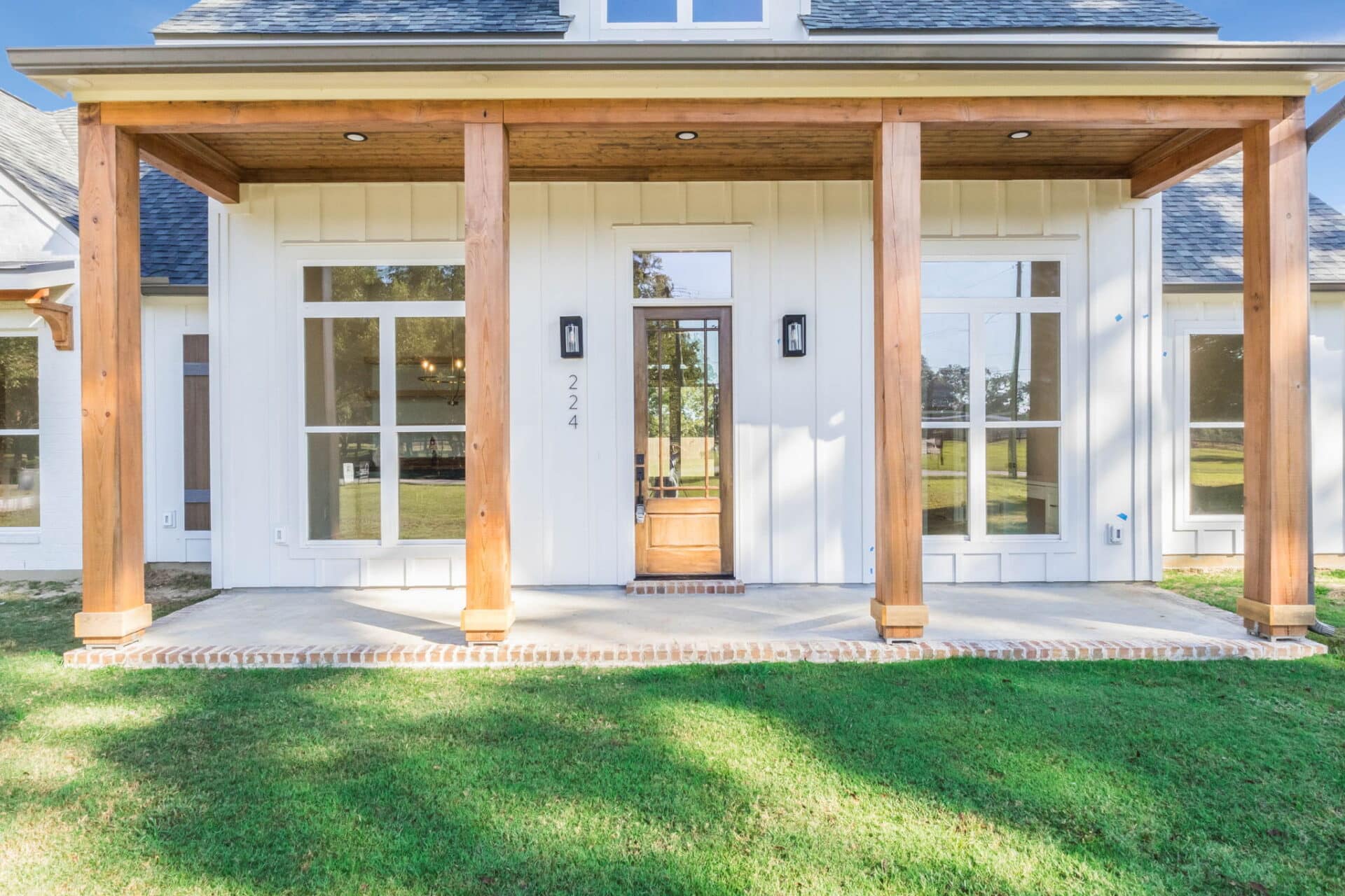 Front view of a house with wood columns, white siding, large windows, a wooden door with sidelights, brick steps, and a covered porch, set on a green lawn.