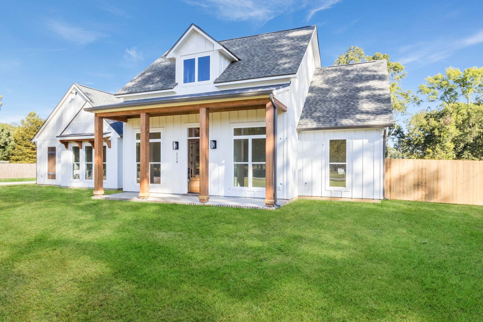 A white modern farmhouse with a gray roof, wood columns on a front porch, large windows, and a well-maintained green lawn under a clear blue sky. A wooden fence and trees are visible in the background.