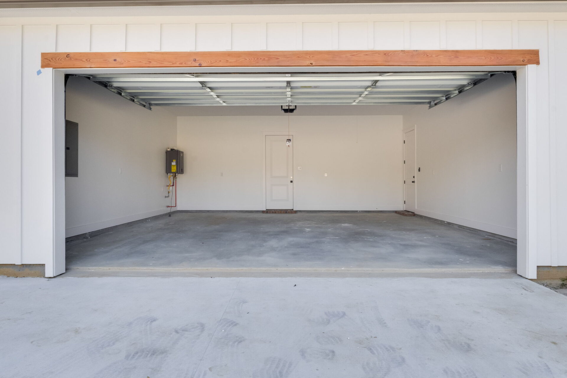 A clean, empty two-car garage with white walls, a concrete floor, and an open garage door. There is a closed door at the back wall and a utility panel mounted on the left side.