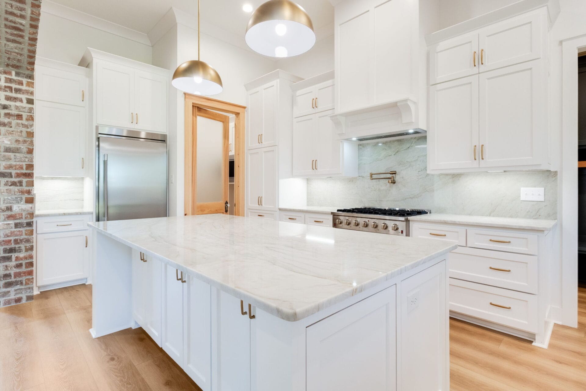 A modern kitchen with white cabinets, a large marble island, stainless steel appliances, brass hardware, two pendant lights, a brick accent wall, and light wood flooring.