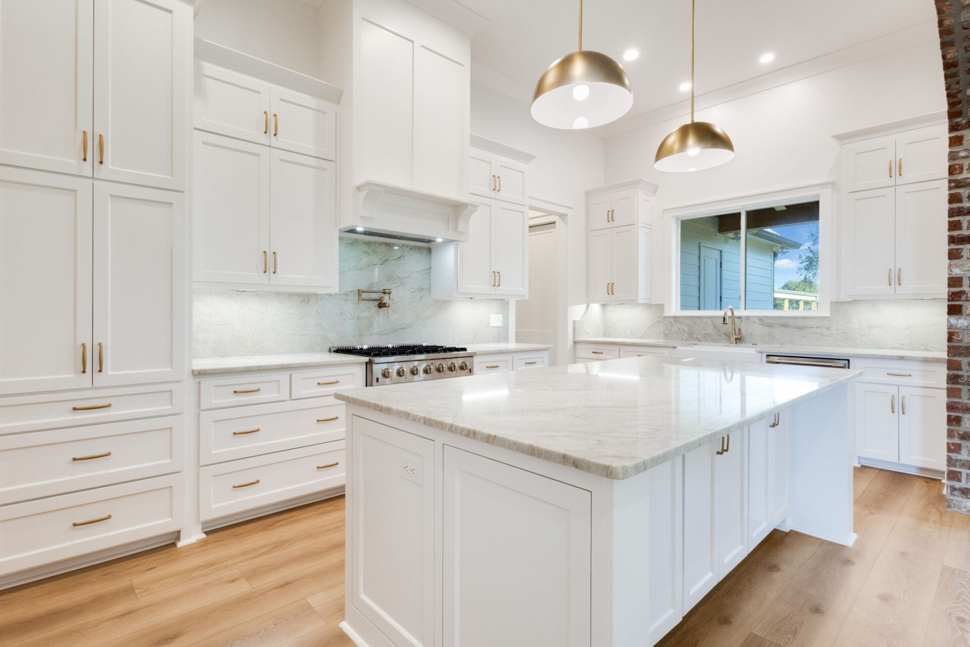 A modern kitchen with white cabinets, a marble island countertop, light wooden floors, stainless steel appliances, two pendant lights, and a large window above the sink.