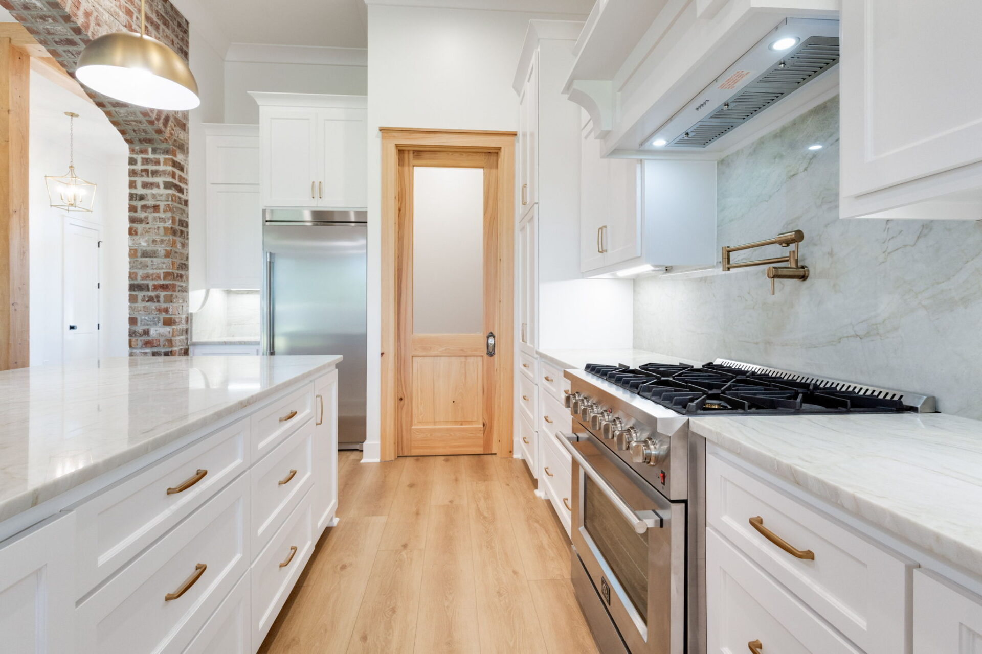 A modern kitchen with white cabinets, marble countertops, a stainless steel gas range, a matching refrigerator, light wood floors, a brick accent wall, and a wooden door with frosted glass.