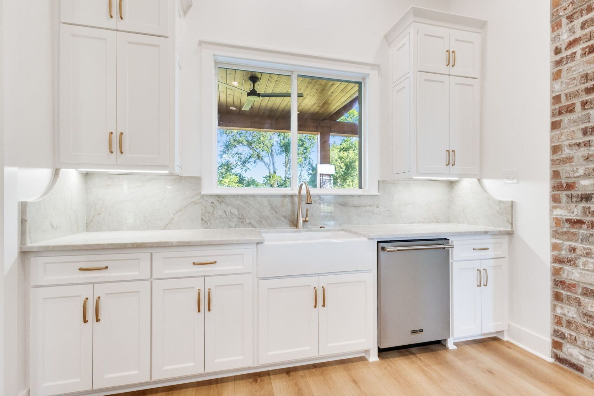 A modern kitchen with white cabinets, gold handles, a large farmhouse sink, marble backsplash and countertops, a small stainless steel dishwasher, light wood flooring, and a window overlooking a porch and trees.