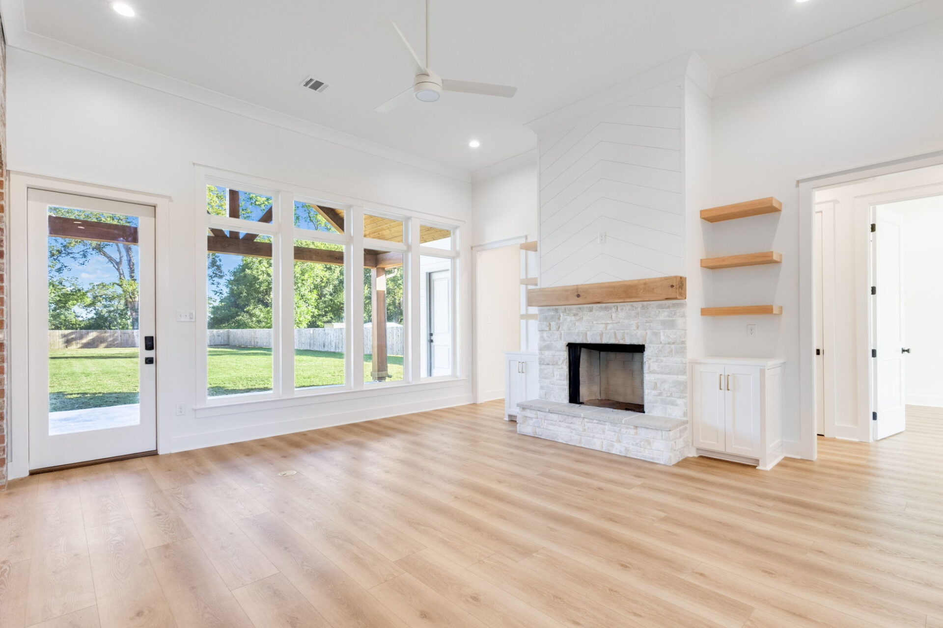 A spacious, empty living room with light wood floors, large windows showing a green yard, a ceiling fan, a white fireplace with a stone base, built-in shelves, and a glass door leading outside.