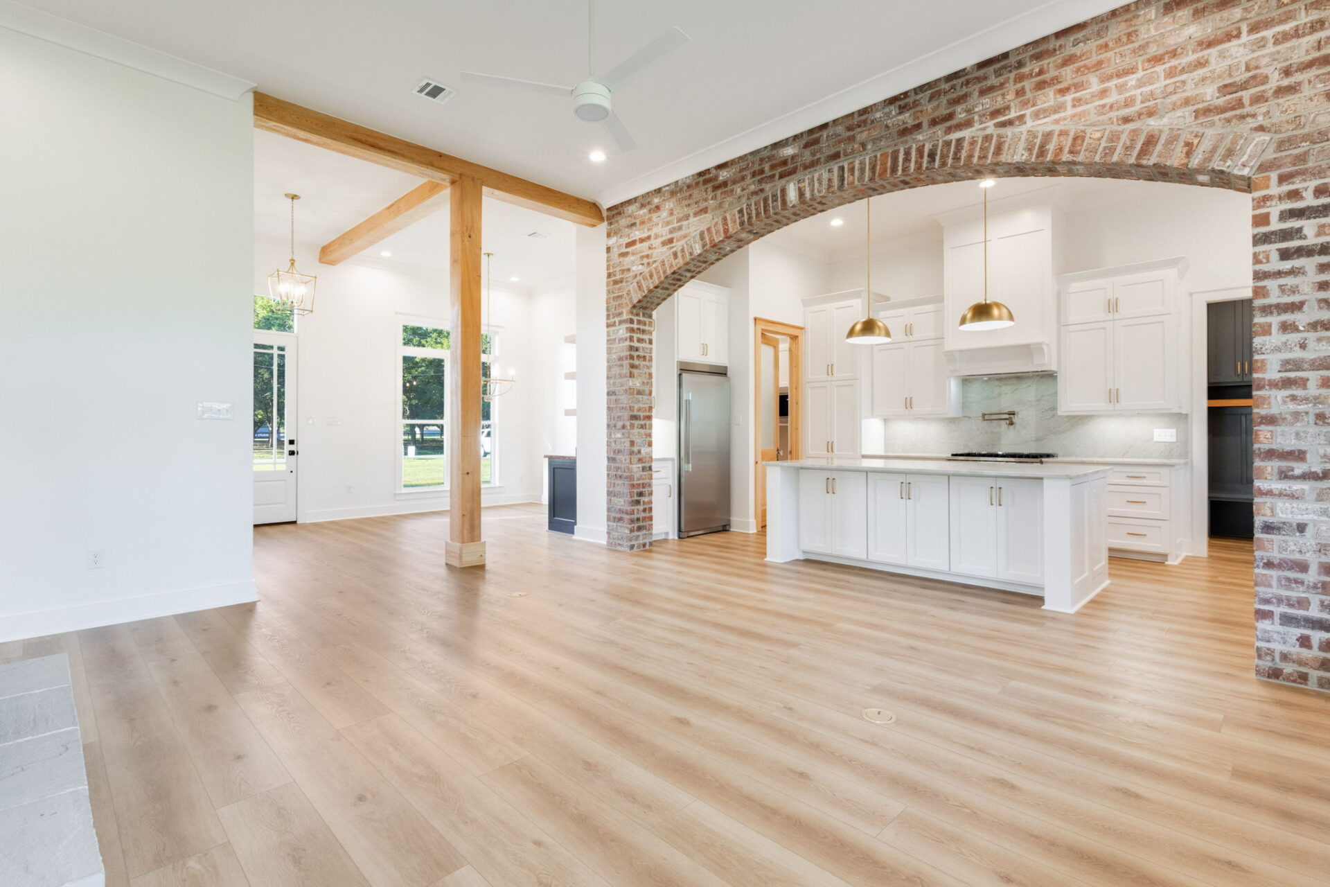 A spacious open-concept kitchen and living area with light wood flooring, exposed brick archway, white cabinets, stainless steel appliances, pendant lights, and large windows letting in natural light.