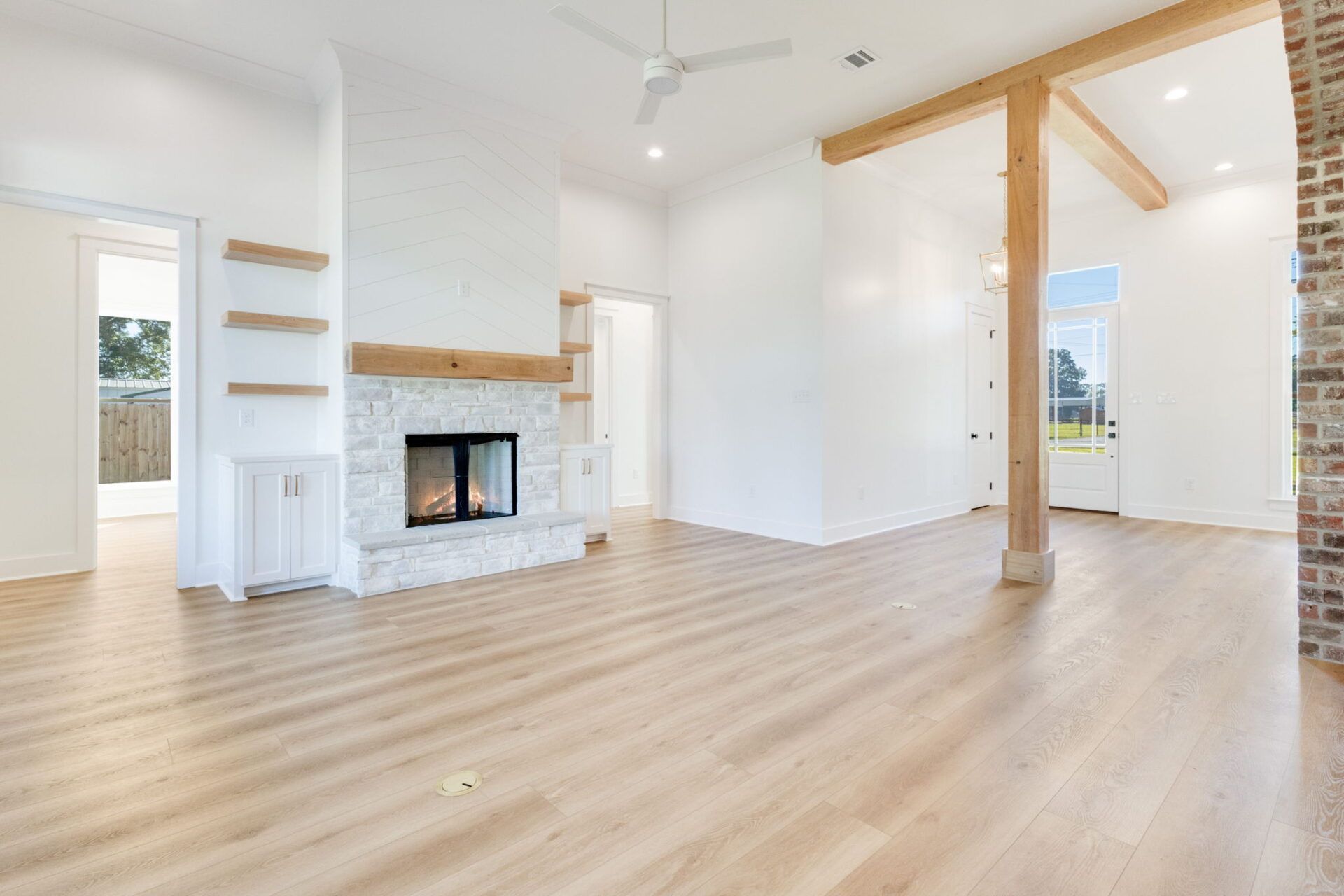 A spacious living room with light wood flooring, a white stone fireplace, floating wood shelves, exposed wooden beams, white walls, and large windows showing an outdoor view.