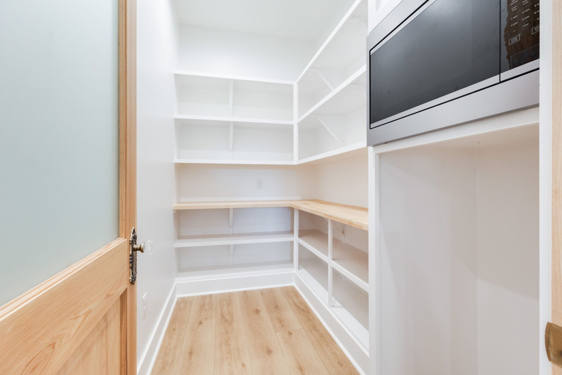 A walk-in pantry with white shelves, light wood countertops, and light wood flooring. A microwave is built into the cabinets on the right, and an open wooden door is on the left. The room is well-lit and empty.