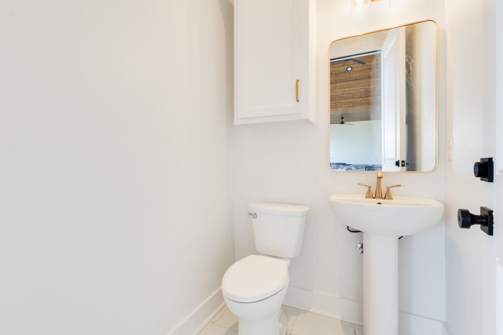 A small bathroom with a white toilet and a white pedestal sink below a rectangular mirror, surrounded by white walls and cabinetry. The faucet and mirror frame are gold-colored.