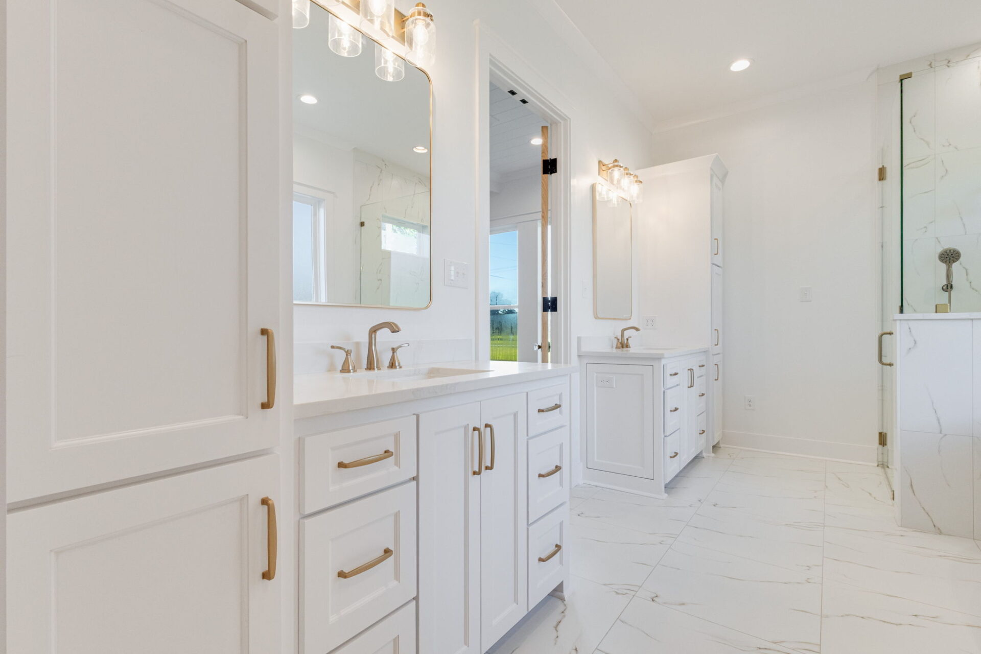 Modern bathroom with white cabinets, gold hardware, dual sinks, large mirrors, marble tile floor, and a glass-enclosed shower with gold fixtures. A window lets in natural light, creating a bright, clean look.