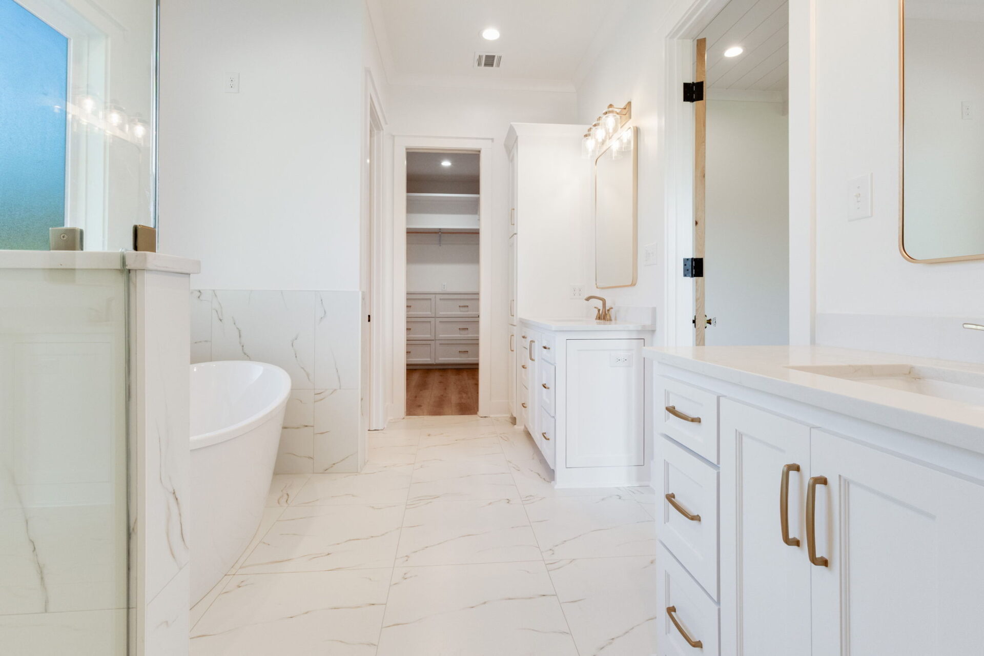 A modern bathroom with white cabinets, tan handles, marble-style tile flooring, a glass shower, a freestanding bathtub, and a large vanity with two sinks; a walk-in closet is visible through an open doorway.