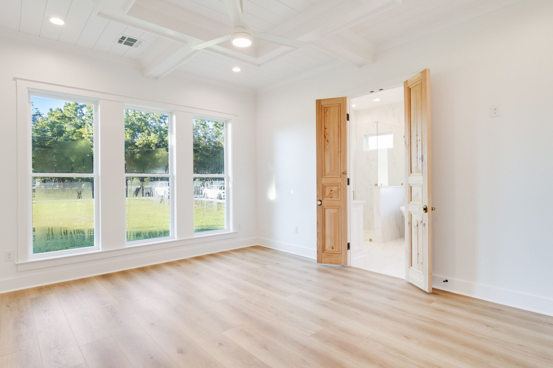 A bright, empty room with light wood flooring, white walls, large windows overlooking greenery, a ceiling fan, and double wooden doors leading to a white-tiled bathroom.