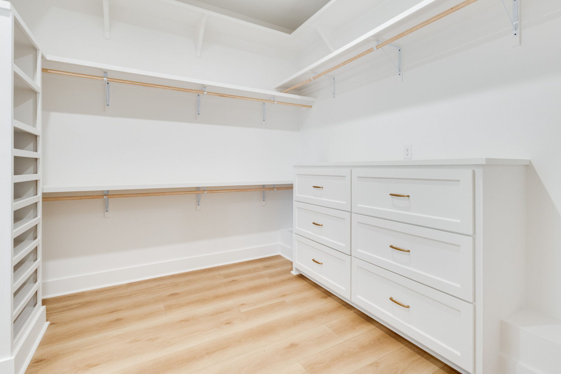 A walk-in closet with white built-in drawers, open shelves, and wooden hanging rods. The floor is light wood, and the walls and trim are white. The space is empty and well-lit.
