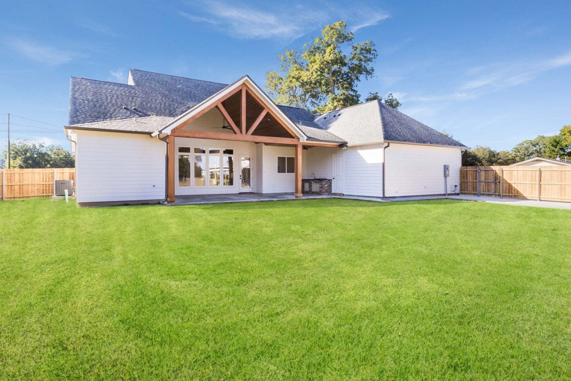 A modern single-story house with white siding, a large covered patio, and a wooden beam accent, situated in a spacious backyard enclosed by a wooden fence under a clear blue sky.
