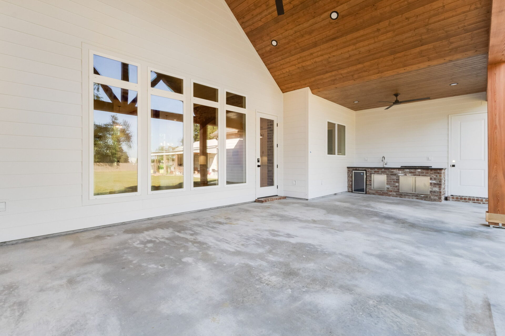 A covered patio with a concrete floor, white paneled walls, large windows, a door, ceiling fans, and a built-in outdoor kitchen area with a grill, sink, and mini fridge.