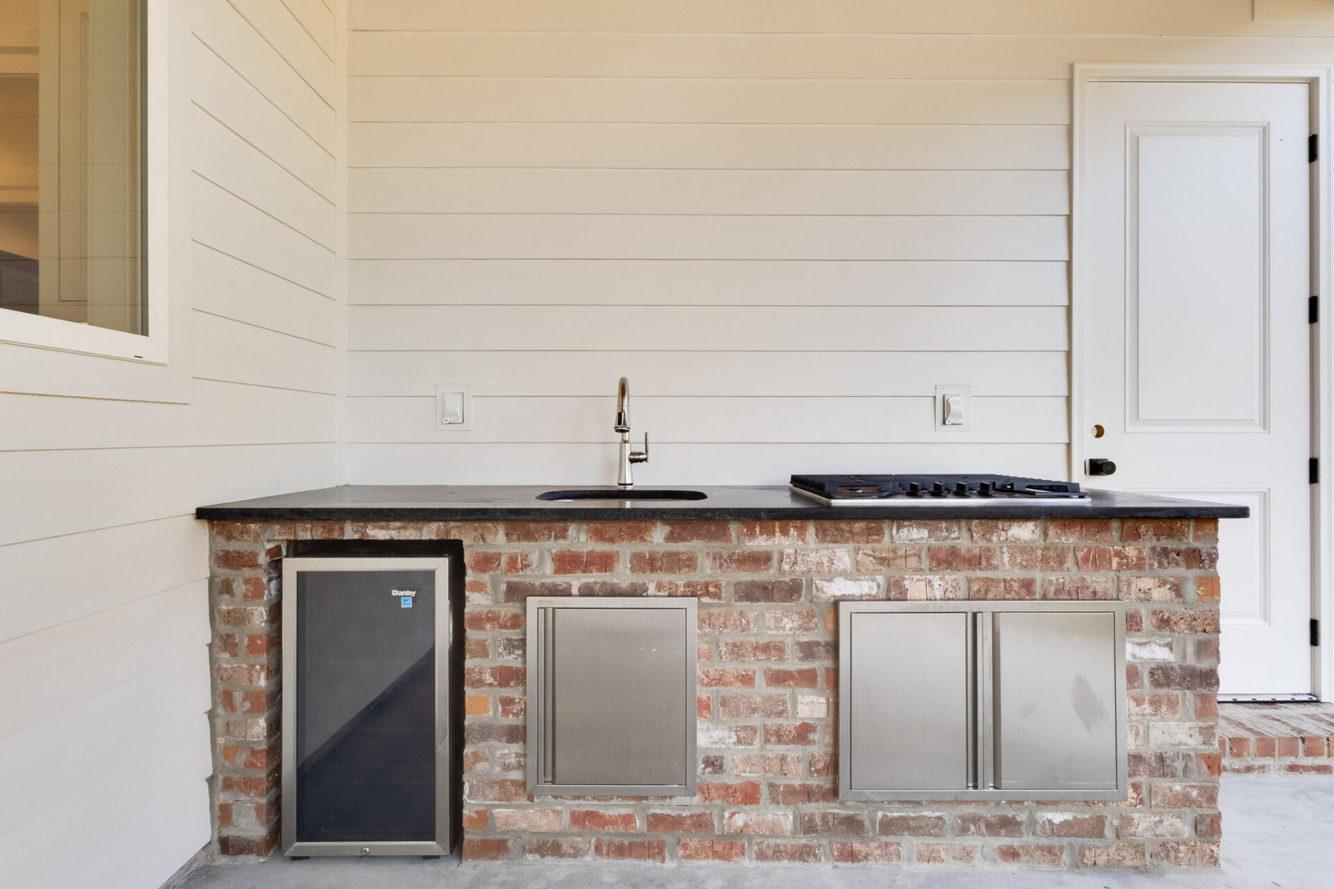 Outdoor kitchen with a brick base featuring a small refrigerator, sink with faucet, black countertop, built-in stovetop, and two stainless steel cabinets, set against a white shiplap wall.