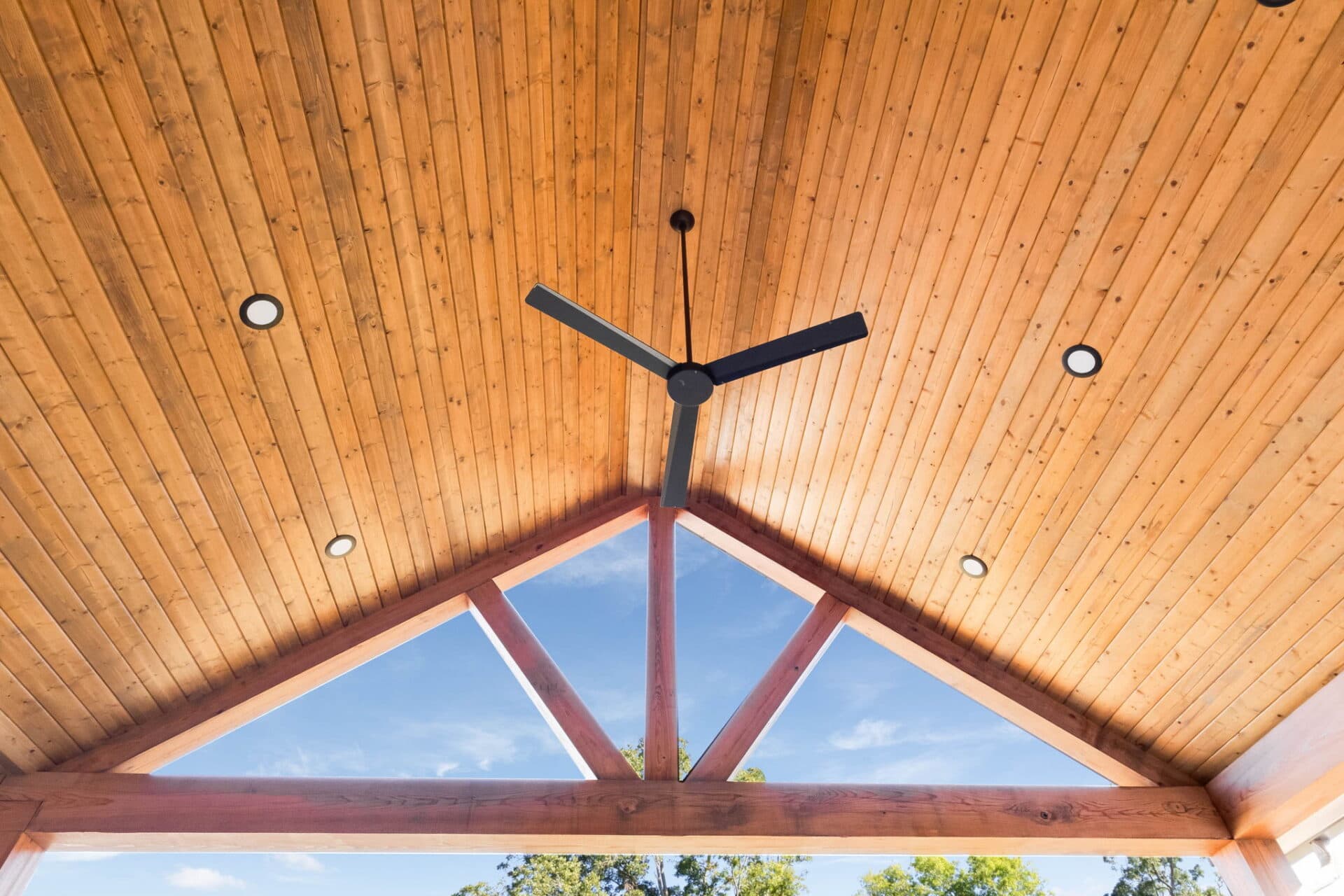 A wooden vaulted ceiling with exposed beams, a black ceiling fan with three blades in the center, and four recessed lights. Blue sky and trees are visible through the open wall below the ceiling.