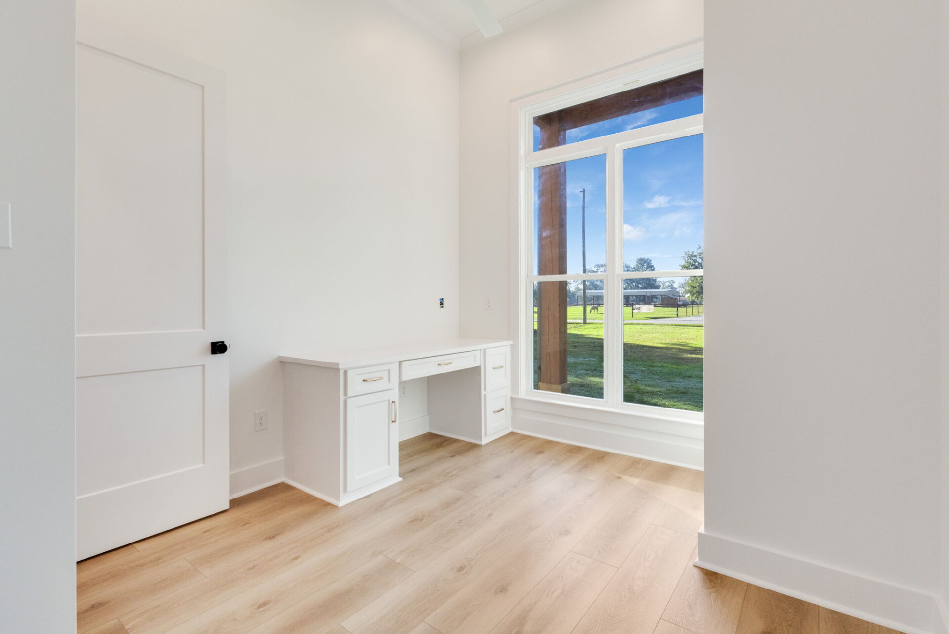 A small, empty room with light wood flooring, white walls, a white built-in desk with drawers, a large window with a view of grass and trees, and a closed white door with a black handle.