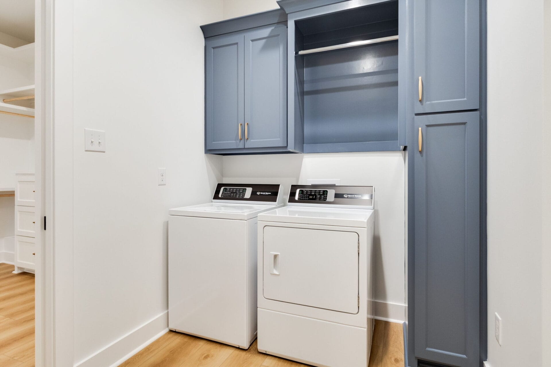 A laundry room with a white washing machine and dryer side by side, under blue cabinets and a hanging rod. The floor is light wood, and the walls are white. An adjacent room is partially visible on the left.