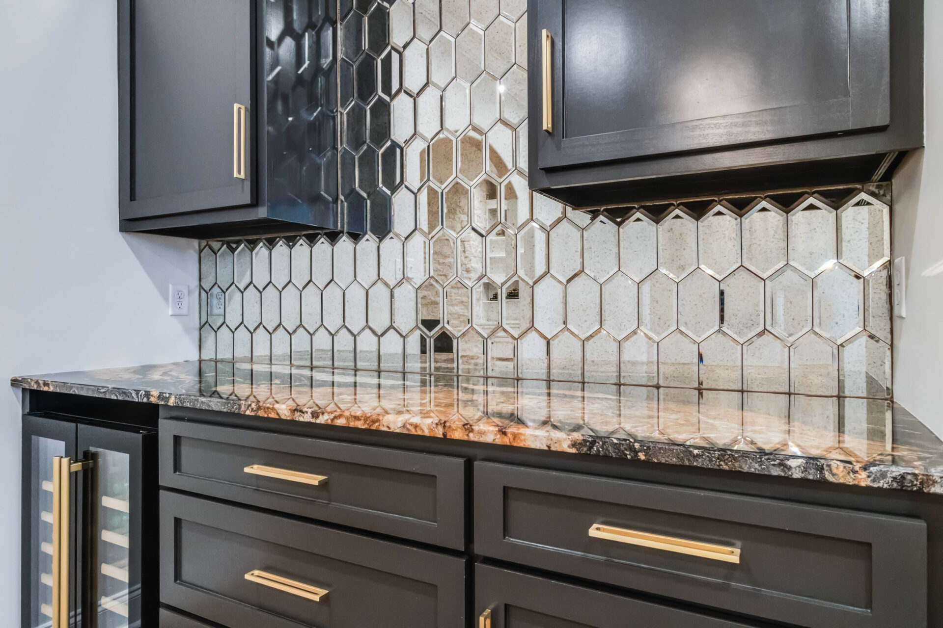 A modern Trahan kitchen features dark cabinets with gold handles, a black and brown marble countertop, and a hexagonal mirrored backsplash. One lower cabinet has a glass door revealing shelves inside.