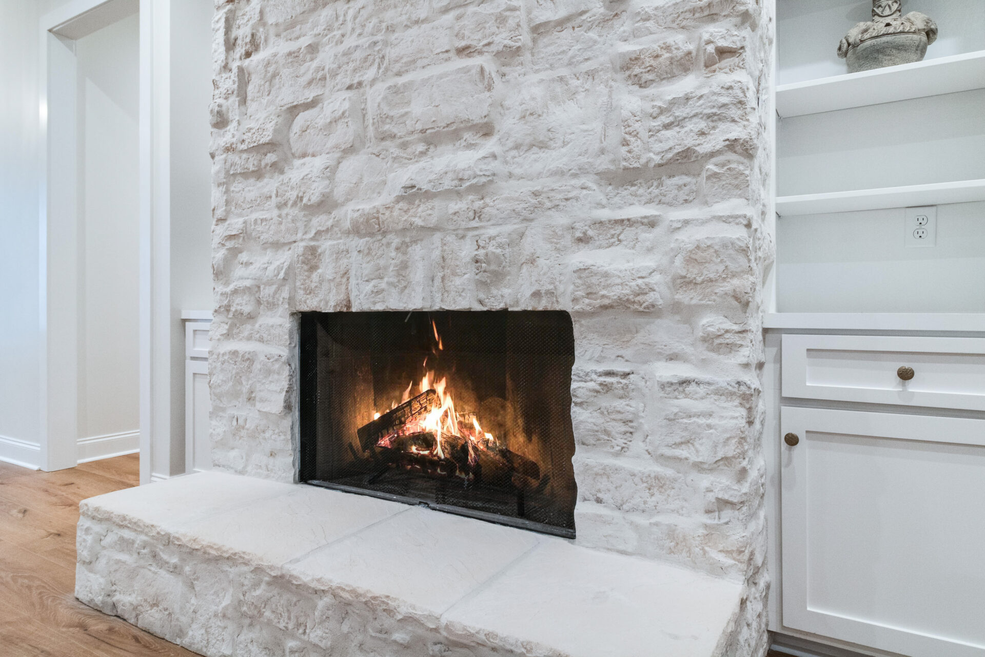 A white stone Trahan fireplace with a lit fire, showing burning logs and flames. Built-in shelving units with cabinets are on the right side of the image. The floor is wooden, and the wall around the fireplace is painted white.