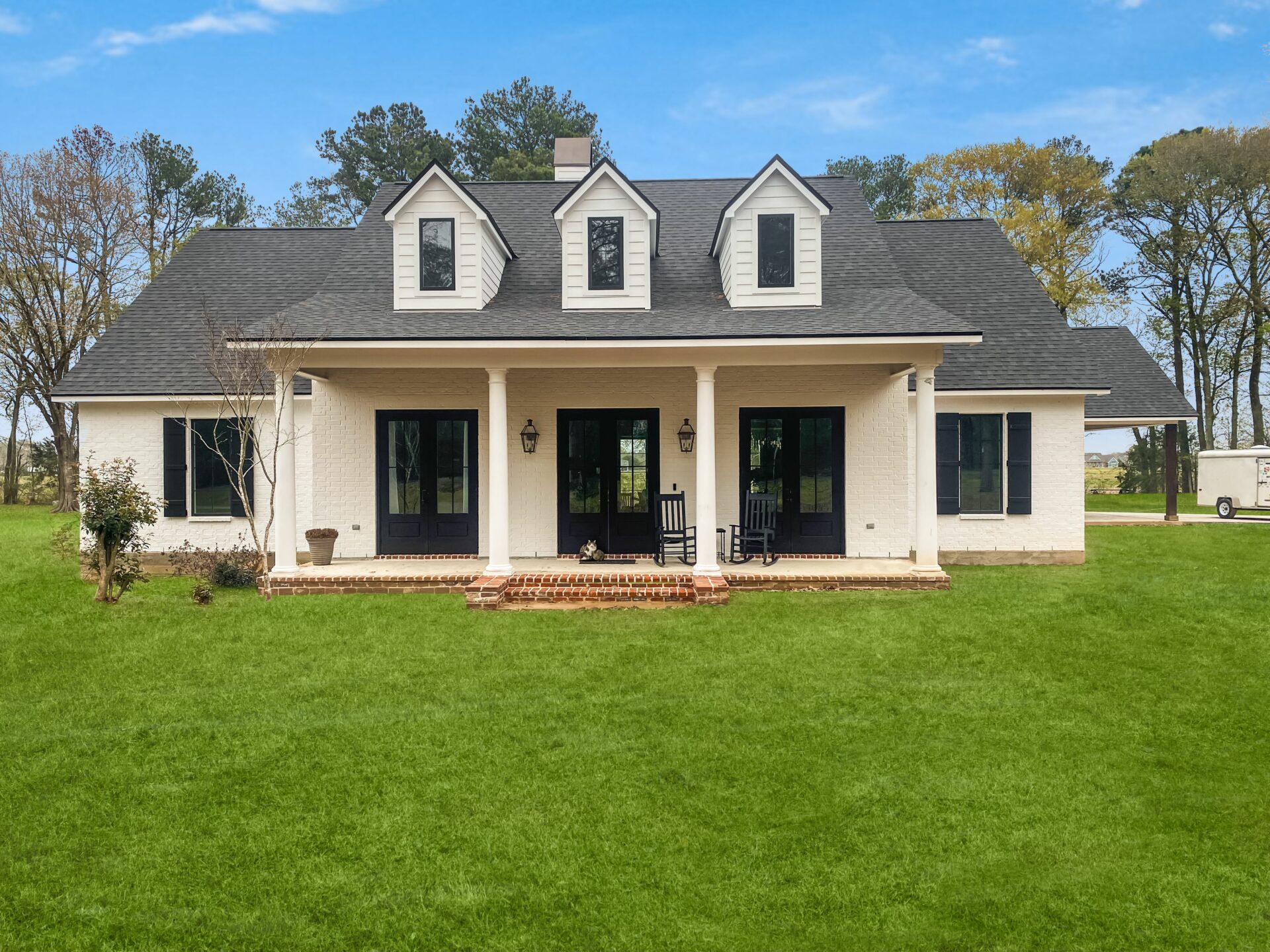 A two-story Trahan house with a gray roof, white exterior, and black window shutters. The house features a covered front porch with two rocking chairs and a brick step leading to the grassy lawn. Trees and a clear blue sky are visible in the background.