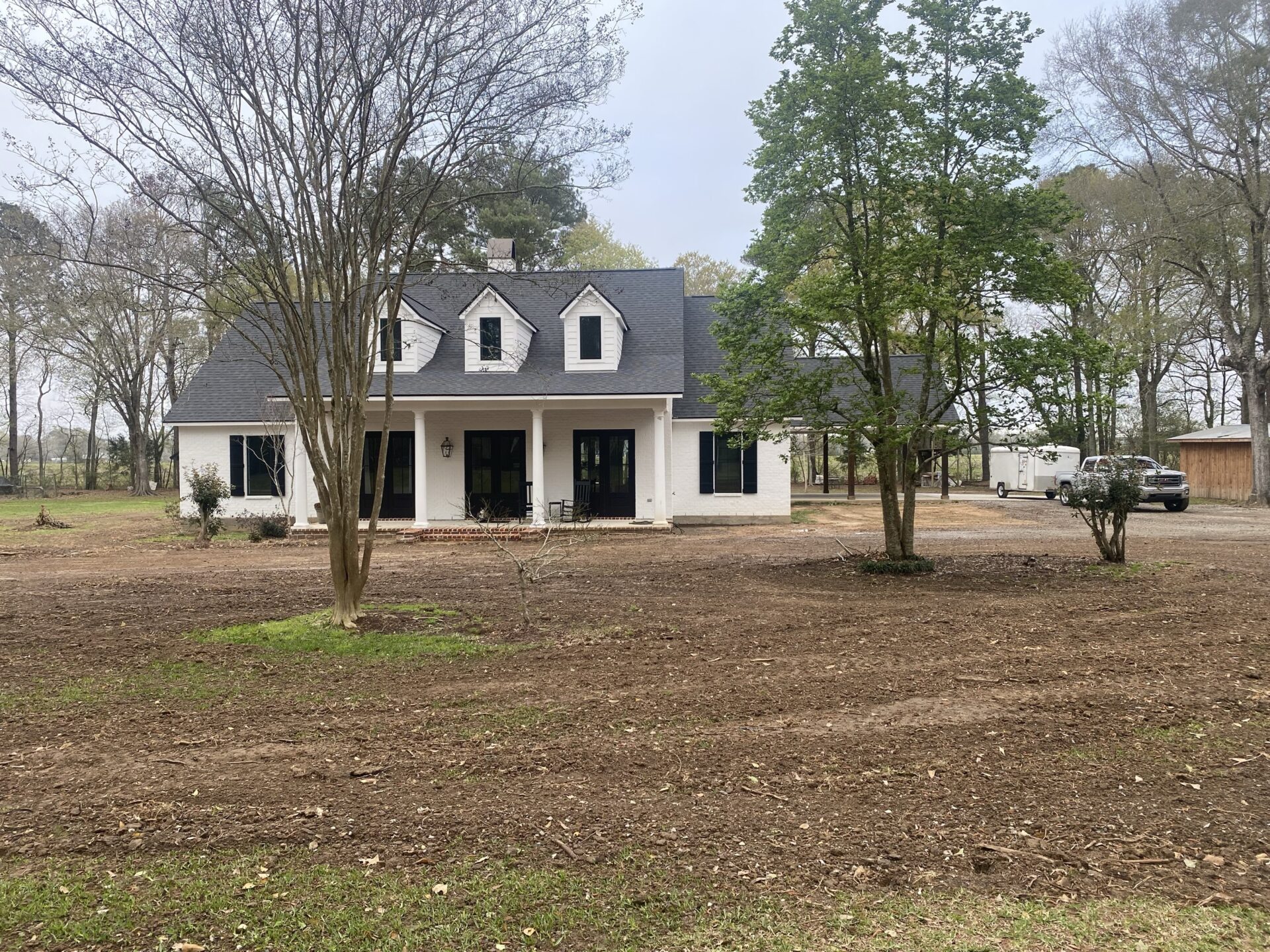 A white, two-story house with a dark gray roof is surrounded by an open lawn and a few trees. The front of the Trahan residence features three dormer windows, two main entrance doors, and a porch light. A vehicle is parked near a detached garage on the right under the cloudy sky.