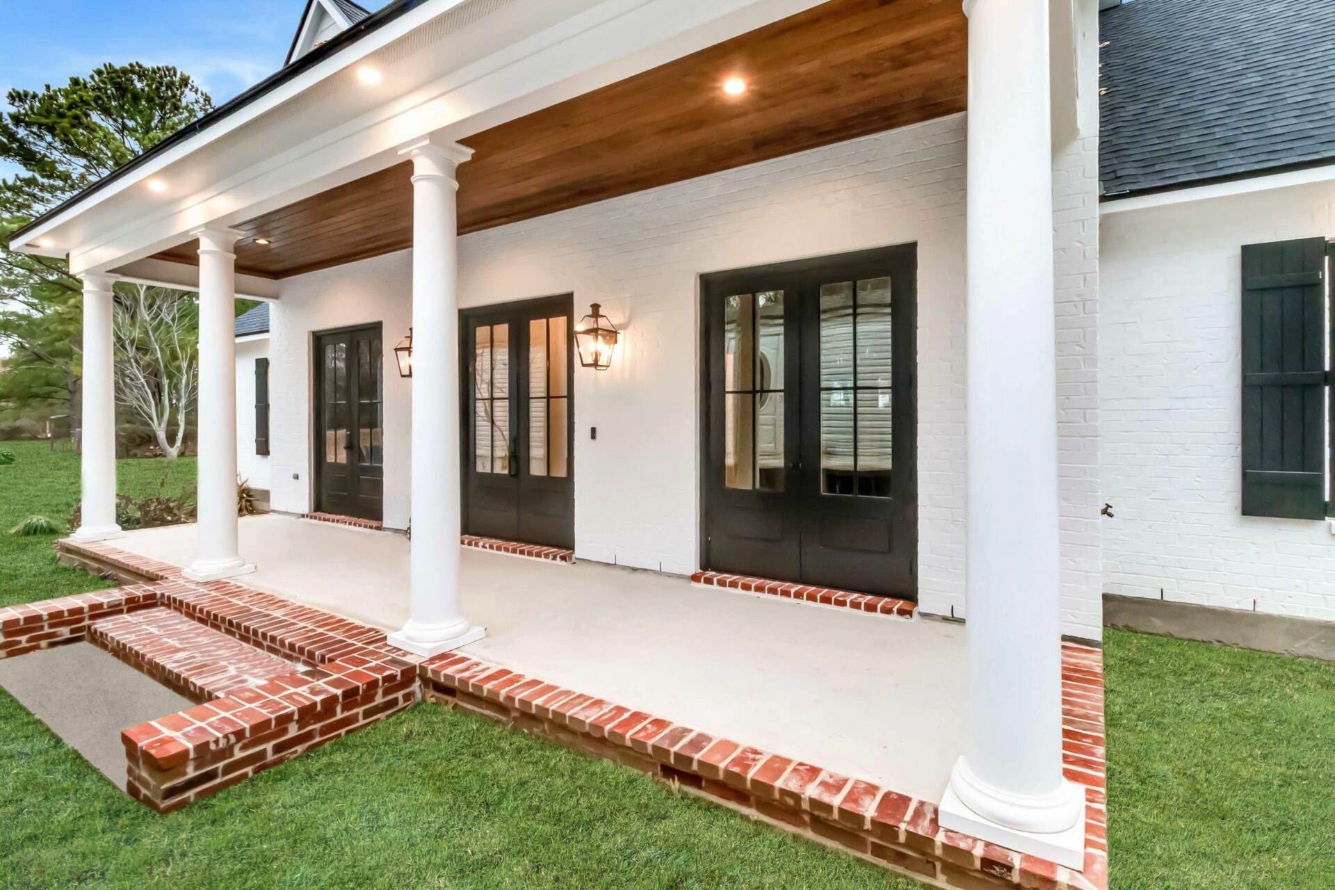 A Trahan-designed house porch features four white columns supporting a wooden ceiling with recessed lighting. The porch has French doors with black frames, red brick steps, and a brick border. The exterior walls are white with a lawn in front.