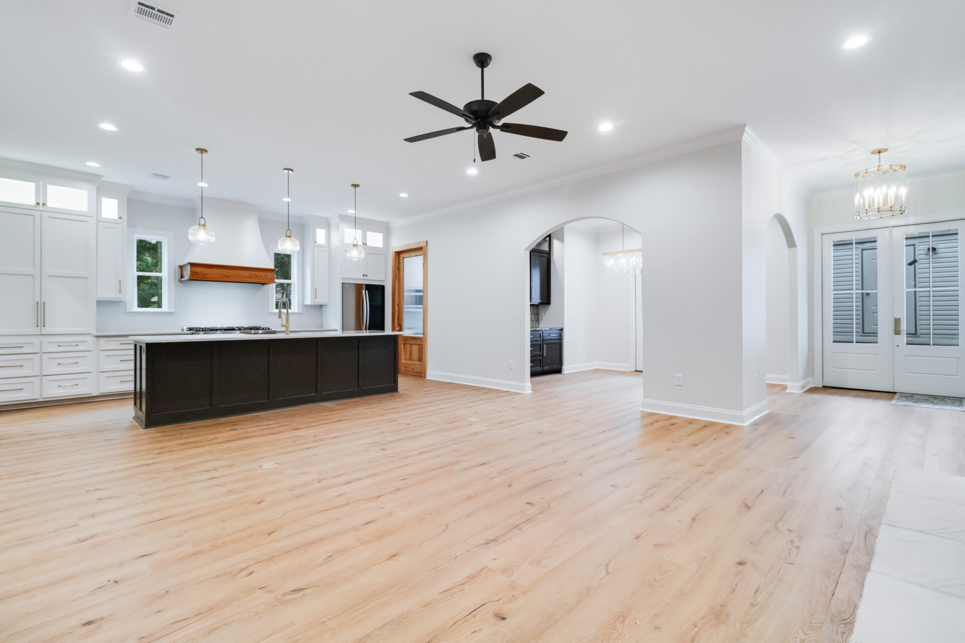 Open-concept living area featuring a spacious kitchen with a large island, white cabinetry, stainless steel appliances, and modern light fixtures. The room has light-colored wood flooring, a ceiling fan, and an arched entryway leading to another room—a signature touch of Trahan designs.