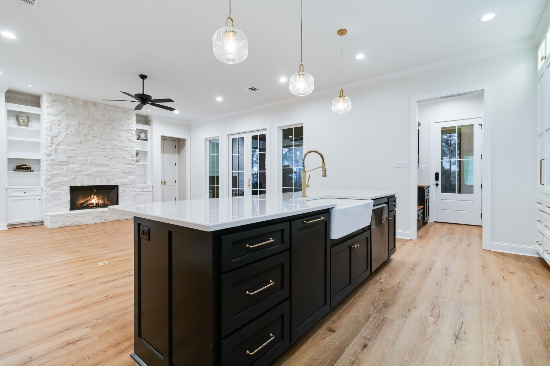 A modern kitchen designed by Trahan features a large island with a white countertop and black cabinetry. Three pendant lights hang above the island. The room has light wood flooring and white walls. In the background, there is a living area with a stone fireplace.