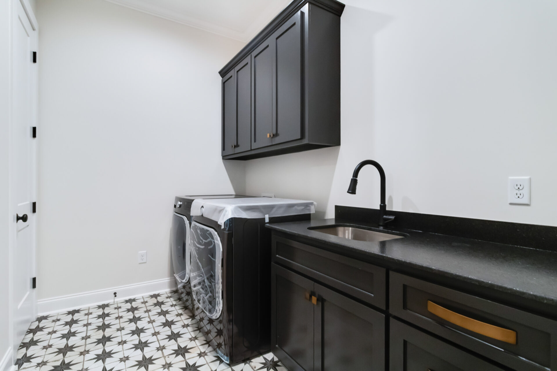 A modern laundry room in the Trahan style features sleek black cabinets and countertops. There is a stainless steel sink with a matching black faucet. A washing machine and dryer sit side by side beneath wall-mounted cabinets, all complemented by a patterned tile floor design.