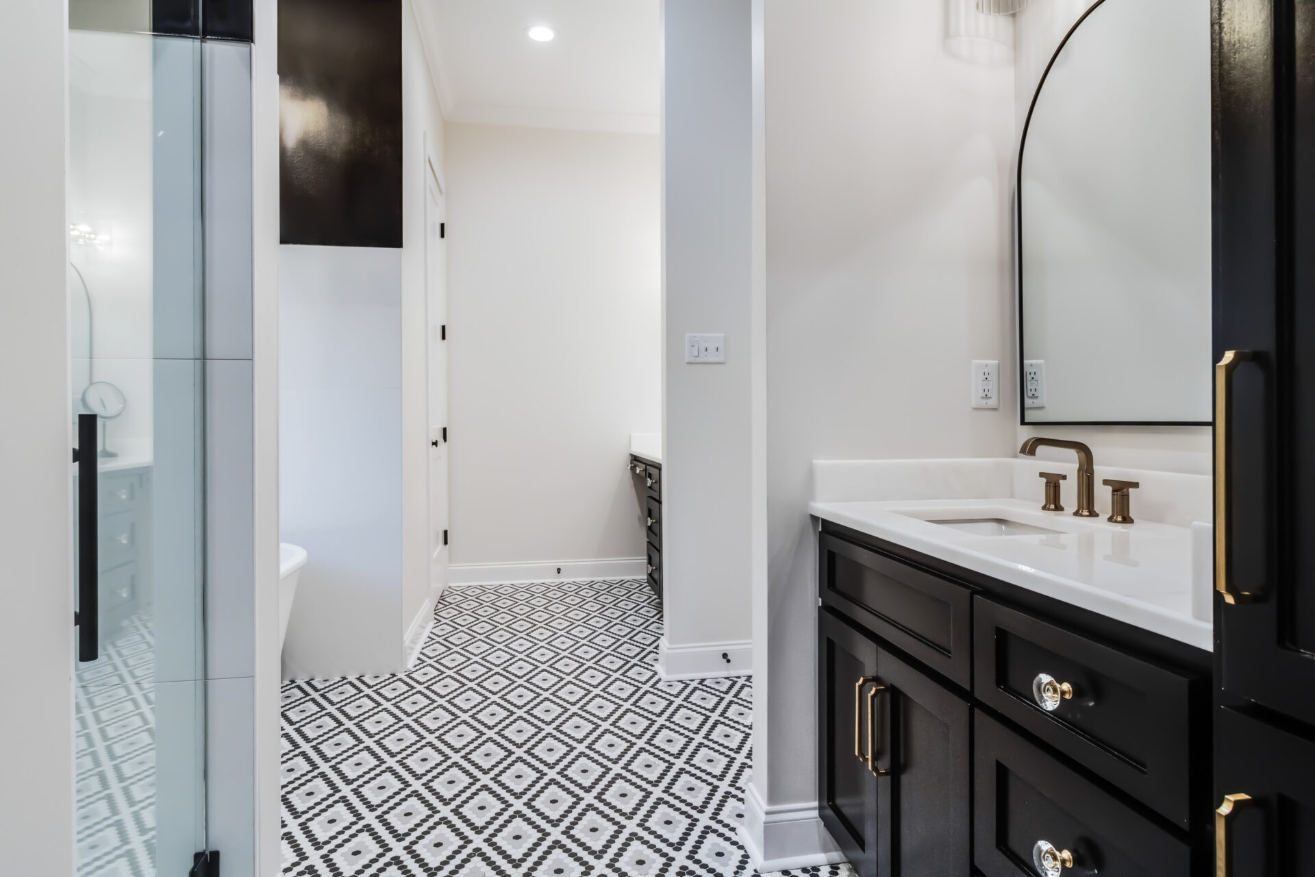 A modern bathroom by Trahan features a black and white geometric tile floor, a double sink vanity with black cabinets and gold hardware, a white quartz countertop, and mounted mirrors. The room is well-lit with both natural and artificial lighting.