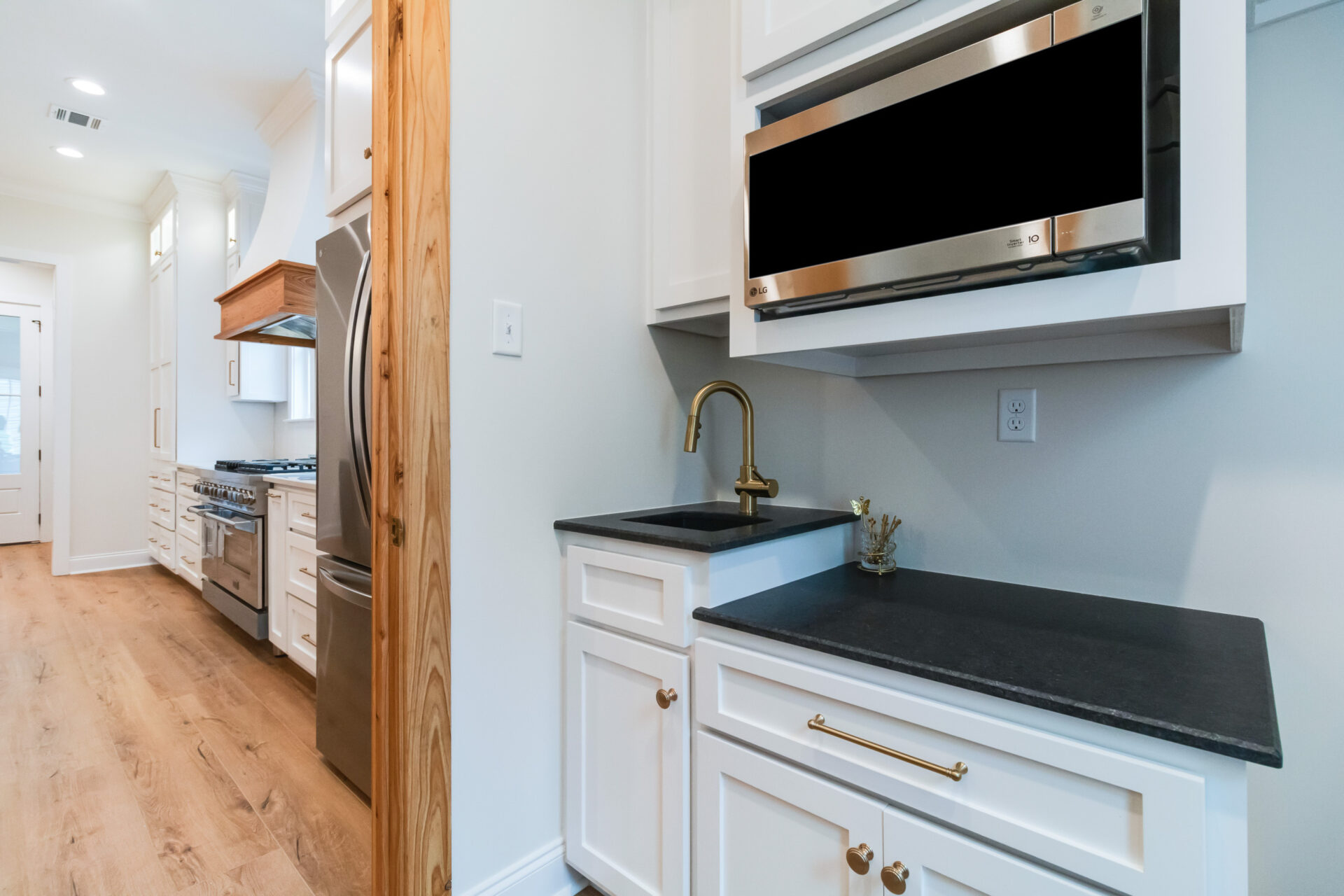A modern Trahan kitchen with white cabinets, black countertops, and wooden flooring. It features a built-in microwave, stainless steel sink with a gold faucet, and a stainless steel oven and refrigerator. The room is well-lit with recessed lighting.